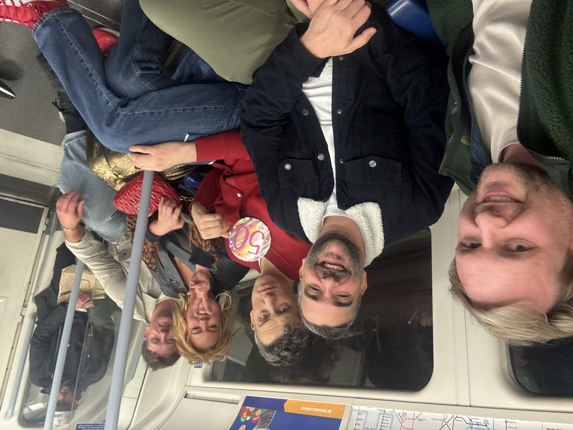 The image shows a group of seven people on a London Underground train. The setting is a typical London Underground carriage interior, with visible handrails, seats, and a route map. The group is arranged across the seating, with some seated and others standing. A woman in a red top holds a "50" badge, suggesting a birthday celebration. The individuals are diverse in age and clothing style. Their expressions suggest a celebratory mood, with smiles and relaxed postures.