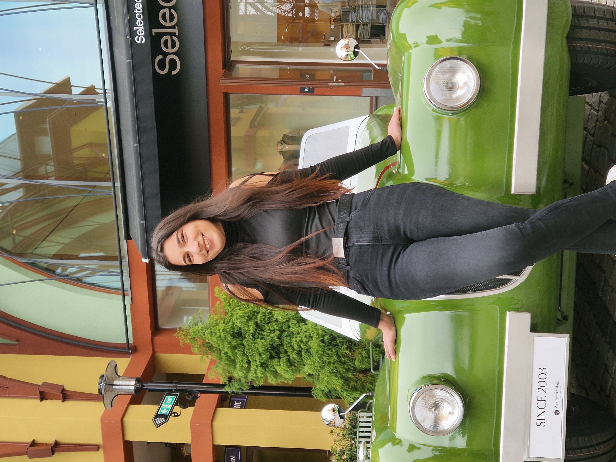 A young adult female is depicted standing outdoors at Wertheim Village, Wertheim, Germany. She is positioned centrally, facing the camera, smiling, and has long, dark brown hair. She wears a black long-sleeved top with cut-out shoulders and dark grey jeans, with her left leg crossed in front of her right. Her hands rest on the hood of a prominent, bright lime green, vintage-style three-wheeled vehicle or sculpture. This vehicle features two circular chrome-bezeled headlights, chrome side mirrors, a small clear windshield, and a chrome strip across its front lower section. A white rectangular sign is attached to the vehicle's front, displaying "SINCE 2003" and "Wertheim Village" with a logo below.
The background consists of modern commercial buildings with yellow and orange/brown facades, incorporating dark brown structural elements. A large glass window reflects parts of the architecture and sky. To the right of the female, a store entrance with a dark awning bears the white lettering "Selected." A mannequin dressed in a jacket is visible through a display window to the right of the vehicle. To the left, a modern street lamp post is present, alongside a green arrow-shaped sign pointing left with "EXIT" written in white. A dense green evergreen shrub is situated behind the vehicle's left side. The ground is paved with cobblestones.