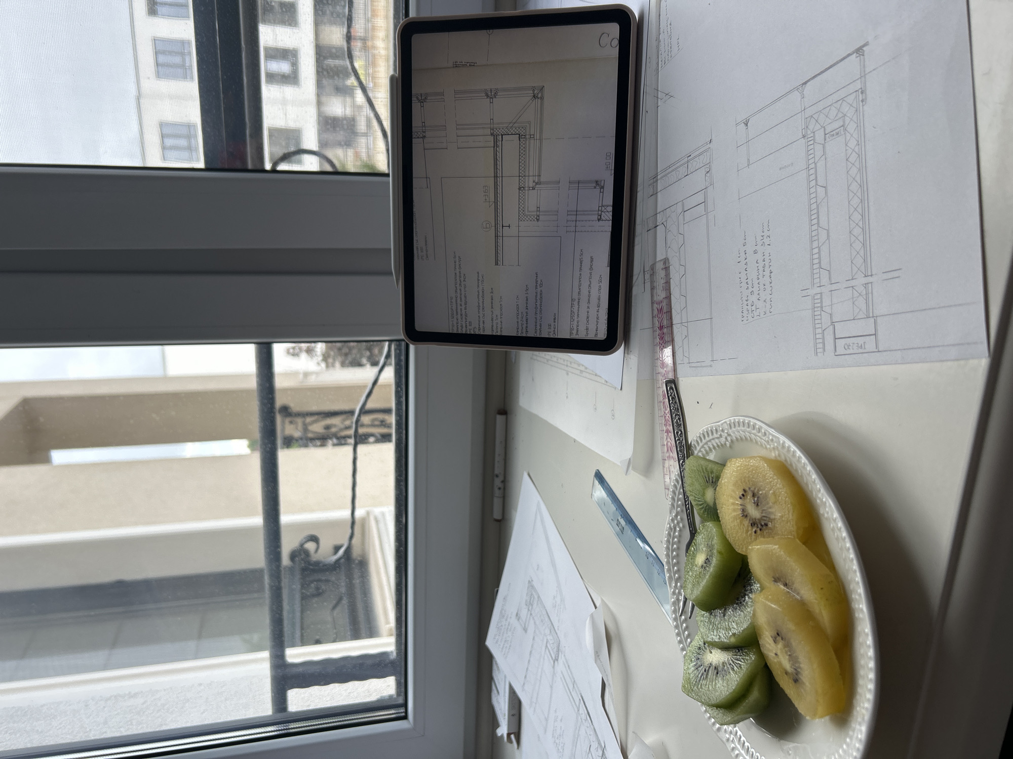 The image shows a desk next to a window, presumably in an interior space. There is a tablet displaying architectural plans, along with physical architectural drawings and tools like a ruler and set square on the desk. A plate of sliced kiwi is also present. Through the window, a view of buildings can be seen. The image does not contain sufficient information to confirm the city, or any information about people.