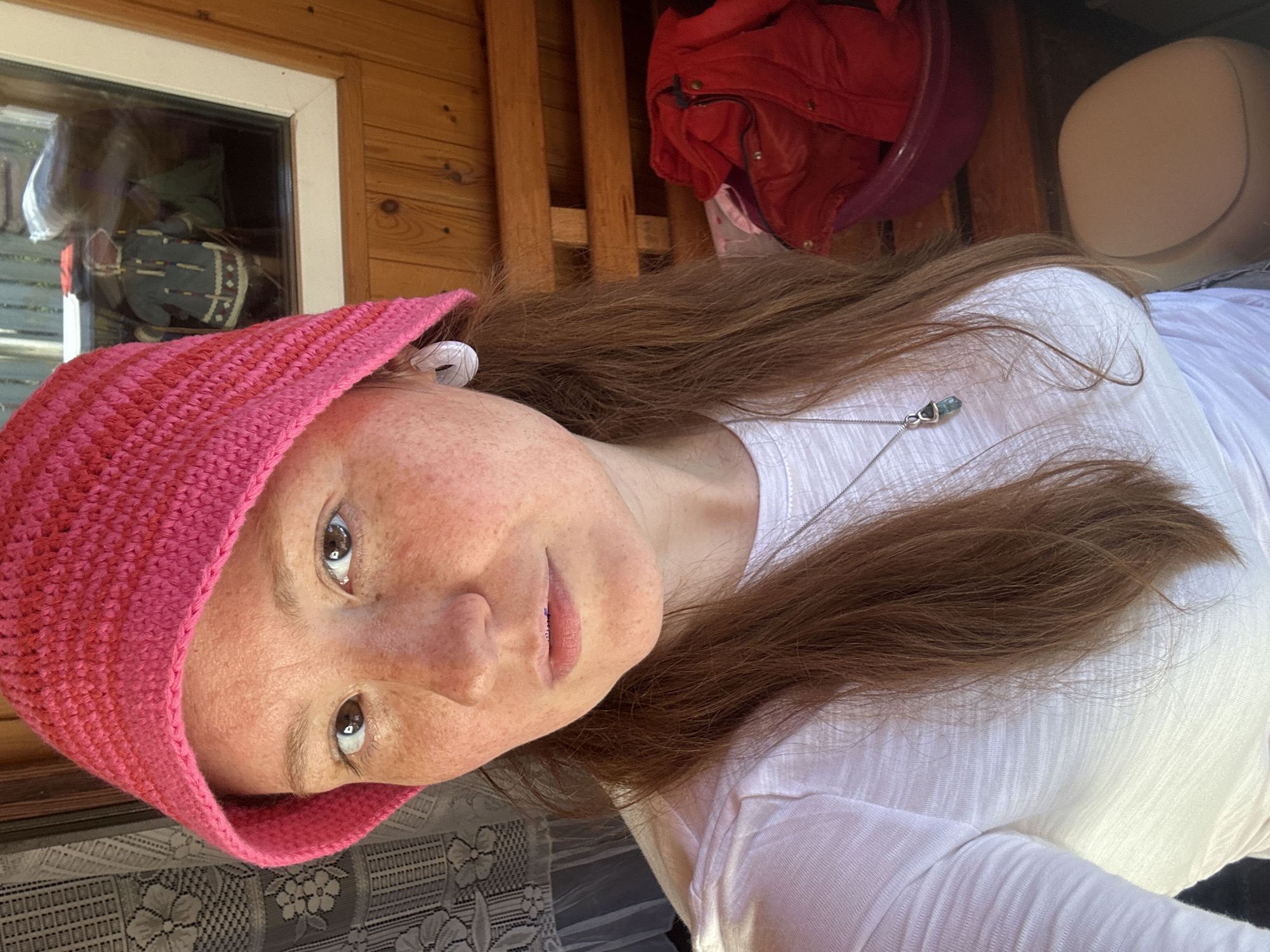 A fair-skinned woman with numerous freckles on her face looks directly at the camera. She has long, reddish-brown hair that falls over her shoulders and wears a vibrant pink knitted or crocheted hat. A silver necklace with a small, light green or blue crystal pendant hangs around her neck, over a white, textured long-sleeved t-shirt.
The background features vertical wooden paneling, suggesting an outdoor or semi-outdoor setting such as a porch or veranda. A window to the left reflects indistinct indoor objects, including a patterned item. To the right, a red jacket is draped over a wooden rail, and a light-colored chair back is partially visible. The natural light indicates it is daytime. The woman's expression is neutral and direct, suggesting she is posing for a photograph. No text is visible in the image. The location is presumably Nizhny Novgorod, Russia.