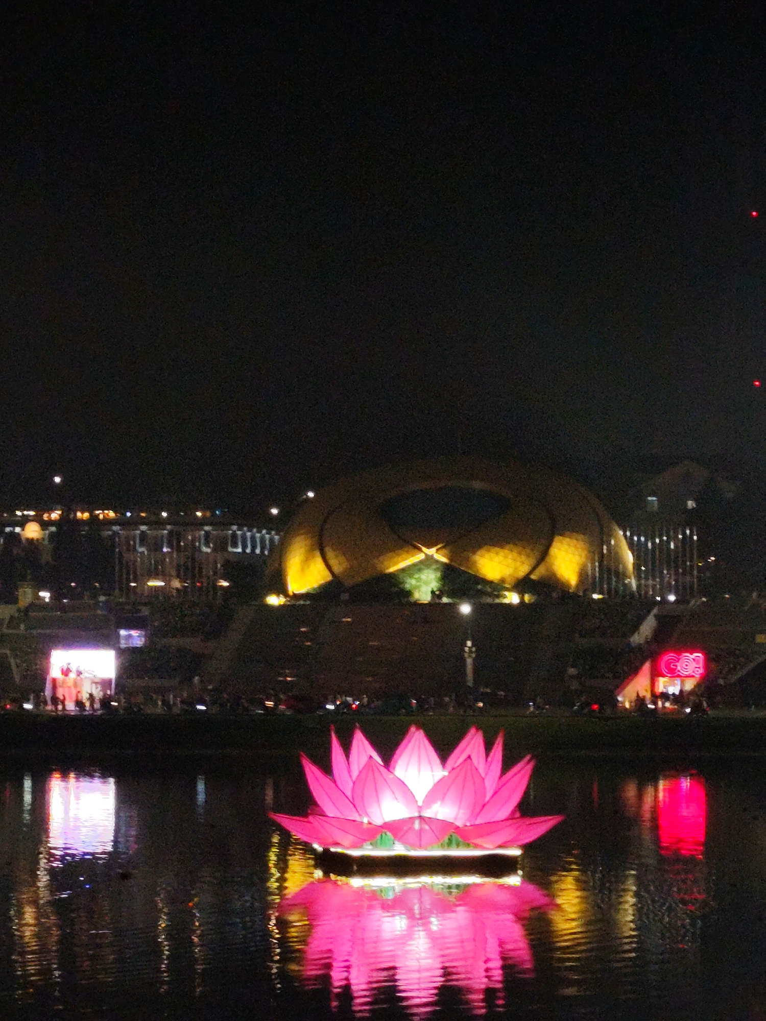 The image is a nighttime view of a lake in Da Lat, Vietnam, with a large, illuminated pink lotus flower floating in the foreground. The flower's reflection is visible in the water. In the background, there is a large, golden-colored dome-shaped structure and several buildings illuminated with lights. A crowd of people is visible on the shore. A sign with the word "Go!" in pink neon is also visible.
