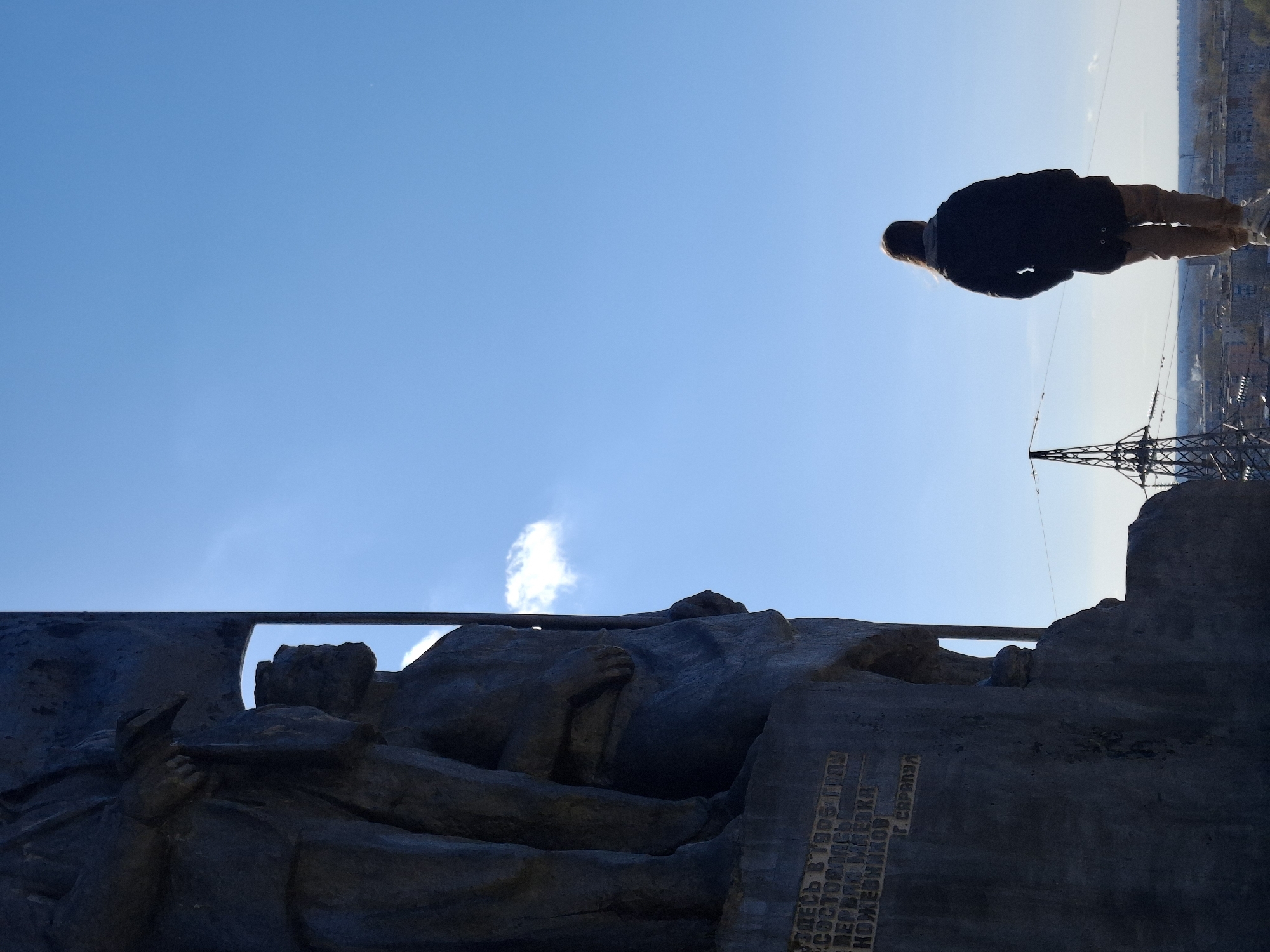 The image depicts a view from below, looking up at a monument and a person standing on a high point. The monument appears to be a stone relief or sculpture depicting figures lying down, with a section of inscription visible in Russian. Above and to the right of the monument, a person wearing a dark jacket and light-colored pants is standing. In the background, a city skyline is faintly visible under a bright sky. Additionally, an electrical tower is partially visible in the background. The monument is located in the city of Sarapul, Russia.