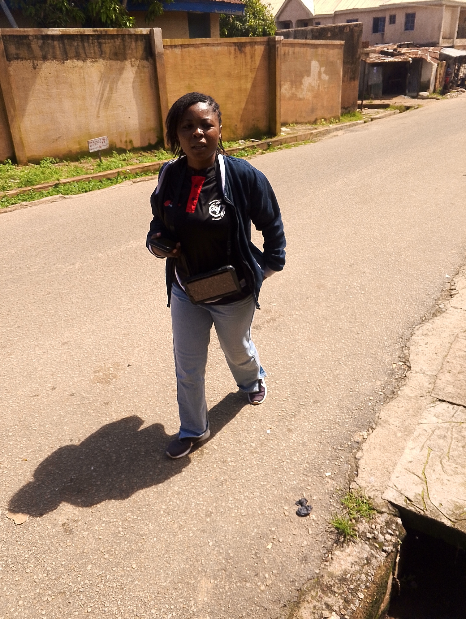 A woman walks along a paved street in an urban area. She is wearing a dark jacket over a black shirt with red accents, blue jeans, and dark sneakers. She has a dark bag slung across her body and is holding a dark object in her right hand. The street is lined with a concrete wall and some vegetation. Buildings are visible in the background. The sun is casting a shadow of the woman on the ground.
