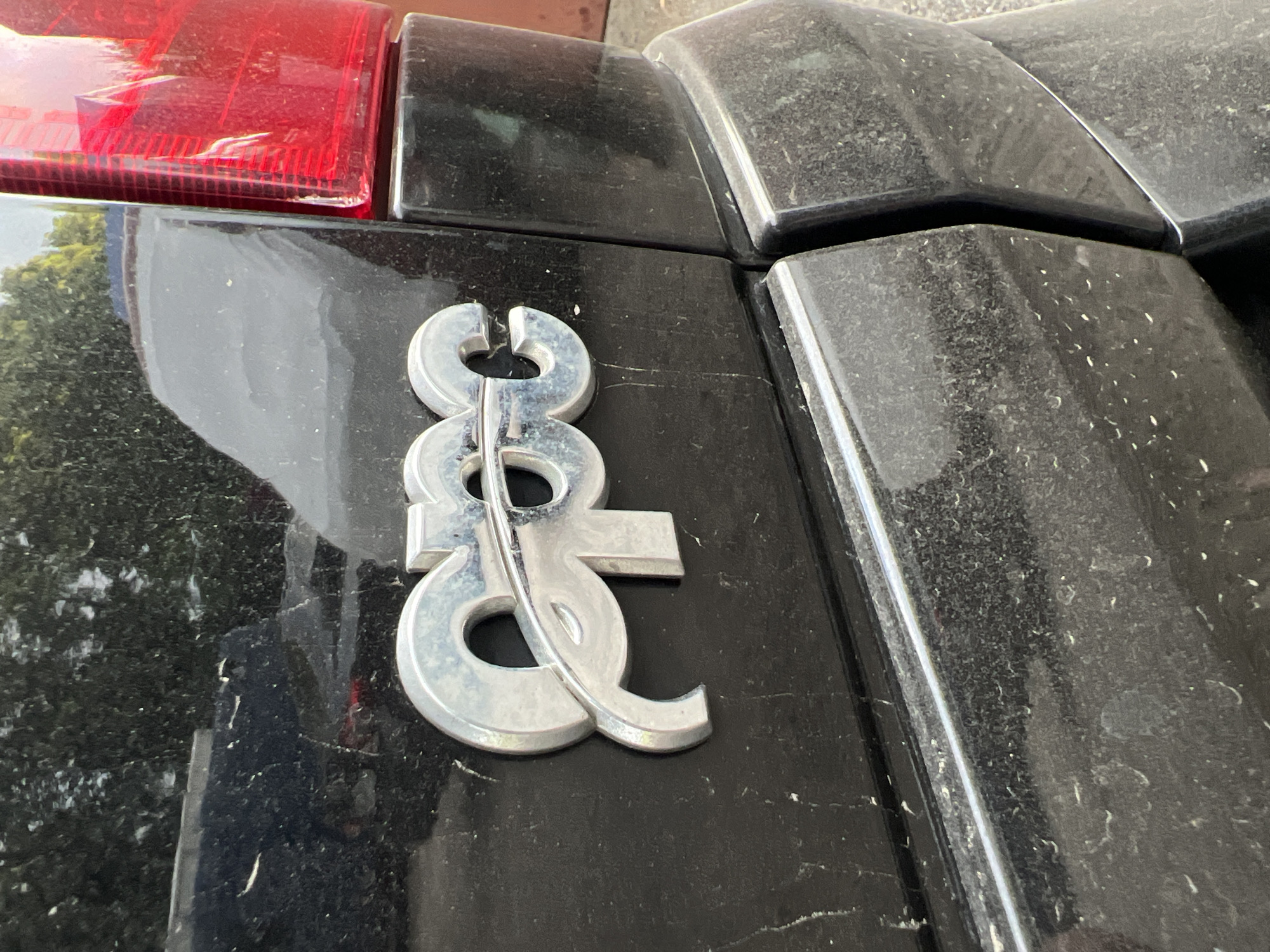 The image is a close-up, eye-level, outdoor shot of a chrome emblem on the rear of a black vehicle. The emblem consists of stylized letters that appear to spell "gpc". The vehicle's surface is covered in a layer of dust. In the background, out of focus, are trees and what appears to be a red taillight of the vehicle. The lighting suggests it is daytime. There are no people visible in the image. The location is unspecified.
