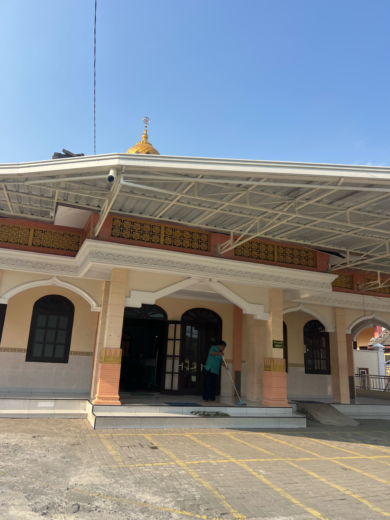 The image shows the exterior of a mosque in Donokerto, Indonesia. A person in a teal shirt and dark pants is sweeping the entrance with a blue mop. The mosque has a light-colored facade with arched windows and doors. Above the entrance, there is a white, sloped roof supported by metal beams. A golden dome topped with a crescent is visible against a clear blue sky. The ground in front of the mosque is paved with interlocking bricks and marked with yellow lines, possibly indicating parking spaces.