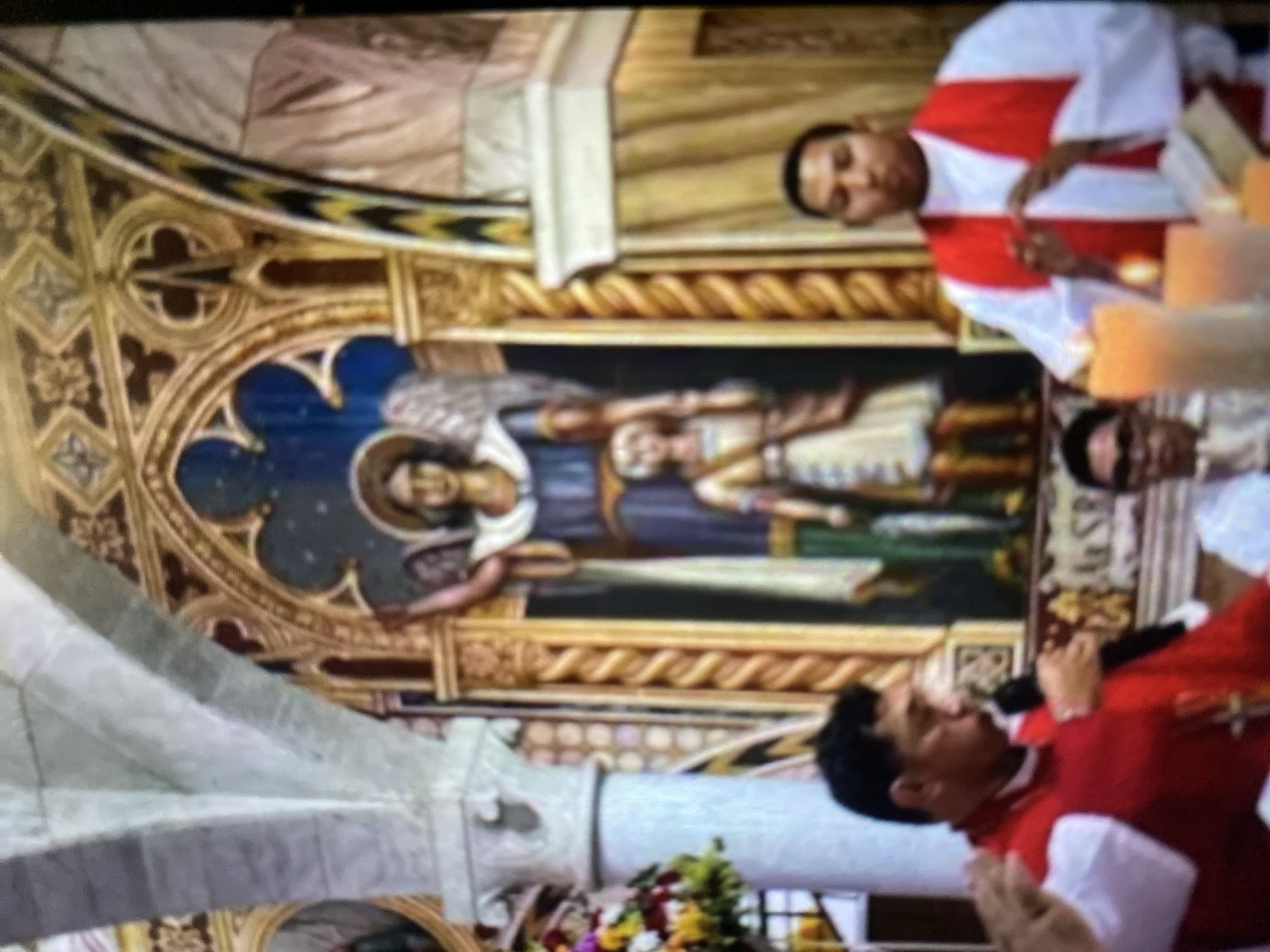 The image appears to be taken inside a church, presumably in Tinaquillo, Venezuela, given the location provided. The church features ornate architecture, including pointed arches, a ribbed vault, and a fresco or painting on the wall depicting figures with wings. Three people are visible, all wearing white shirts and red vestments. One is speaking into a microphone. Another is standing near the painting, while the third is partially visible in the upper right corner. There is also floral decoration.