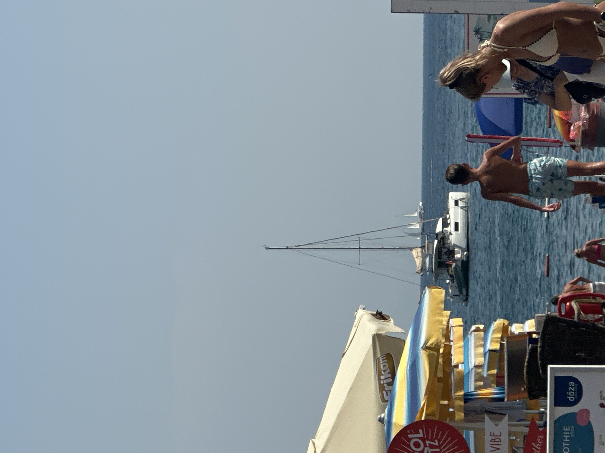 A sunny day at the beach in Boreti, Montenegro features several people and objects. A woman in a white bikini top and patterned dress is bending down, looking at something out of frame. A young boy wearing patterned swim trunks stands facing away from the camera, looking out at the water. Behind them, the calm blue sea is dotted with several sailboats. In the foreground, a collection of colorful beach umbrellas and chairs are set up, with some advertising text visible, including "Frikon" and "doza". There are also signs for "VIBE" and "SMOOTHIE". The scene suggests a relaxed summer atmosphere.