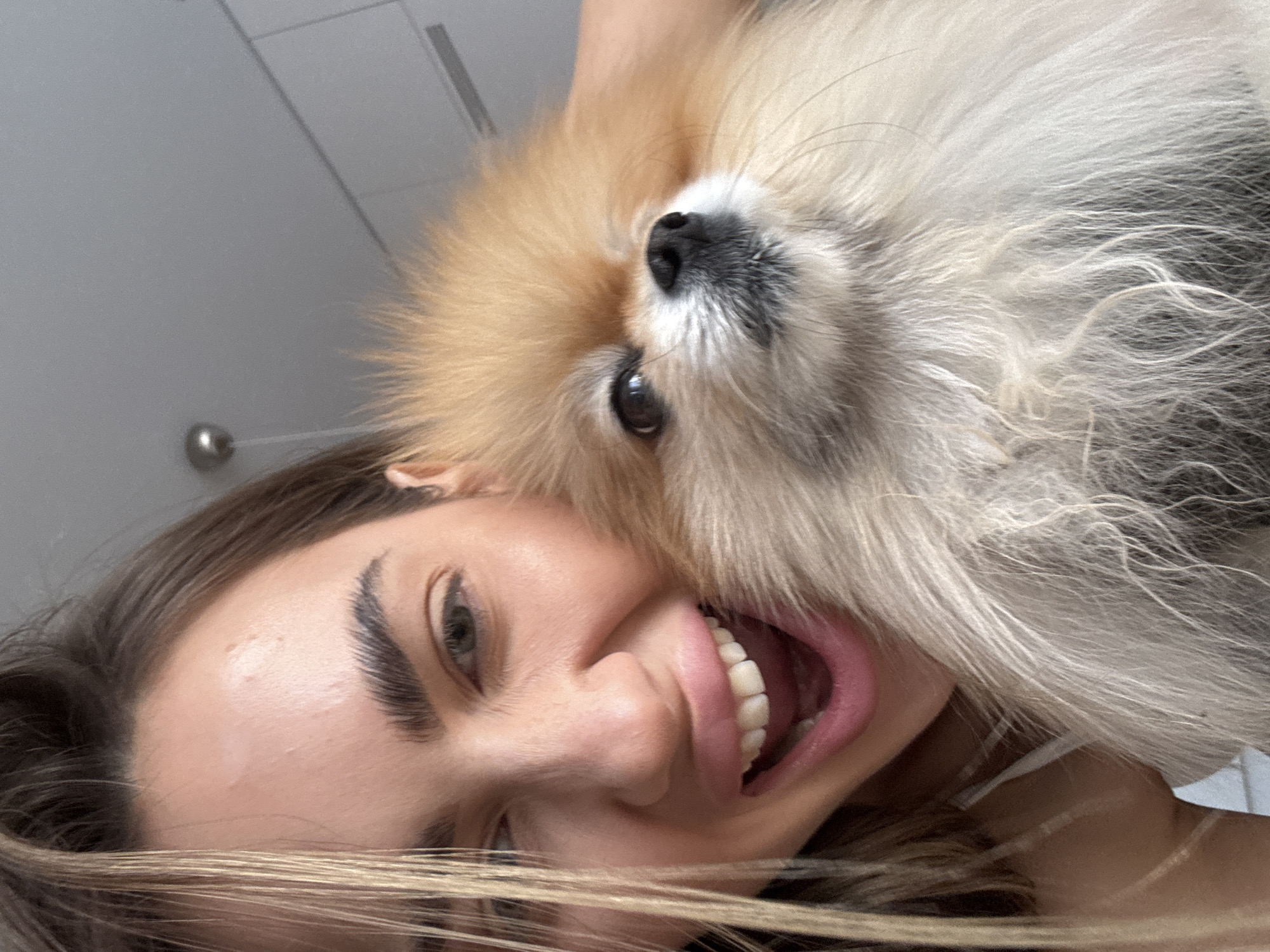A selfie-style photograph captures a woman with her head tilted to the left, positioned alongside a fluffy, light-brown and white dog. The woman is smiling broadly, displaying her teeth. She has fair skin, light-brown hair with blonde highlights, and defined eyebrows. The dog, likely a Pomeranian, has a black nose and dark eyes. The background appears to be a sparsely decorated interior space, possibly a bathroom, with a metallic light fixture visible in the upper left corner. The image provides no discernible information to link the scene to Warsaw, Poland.