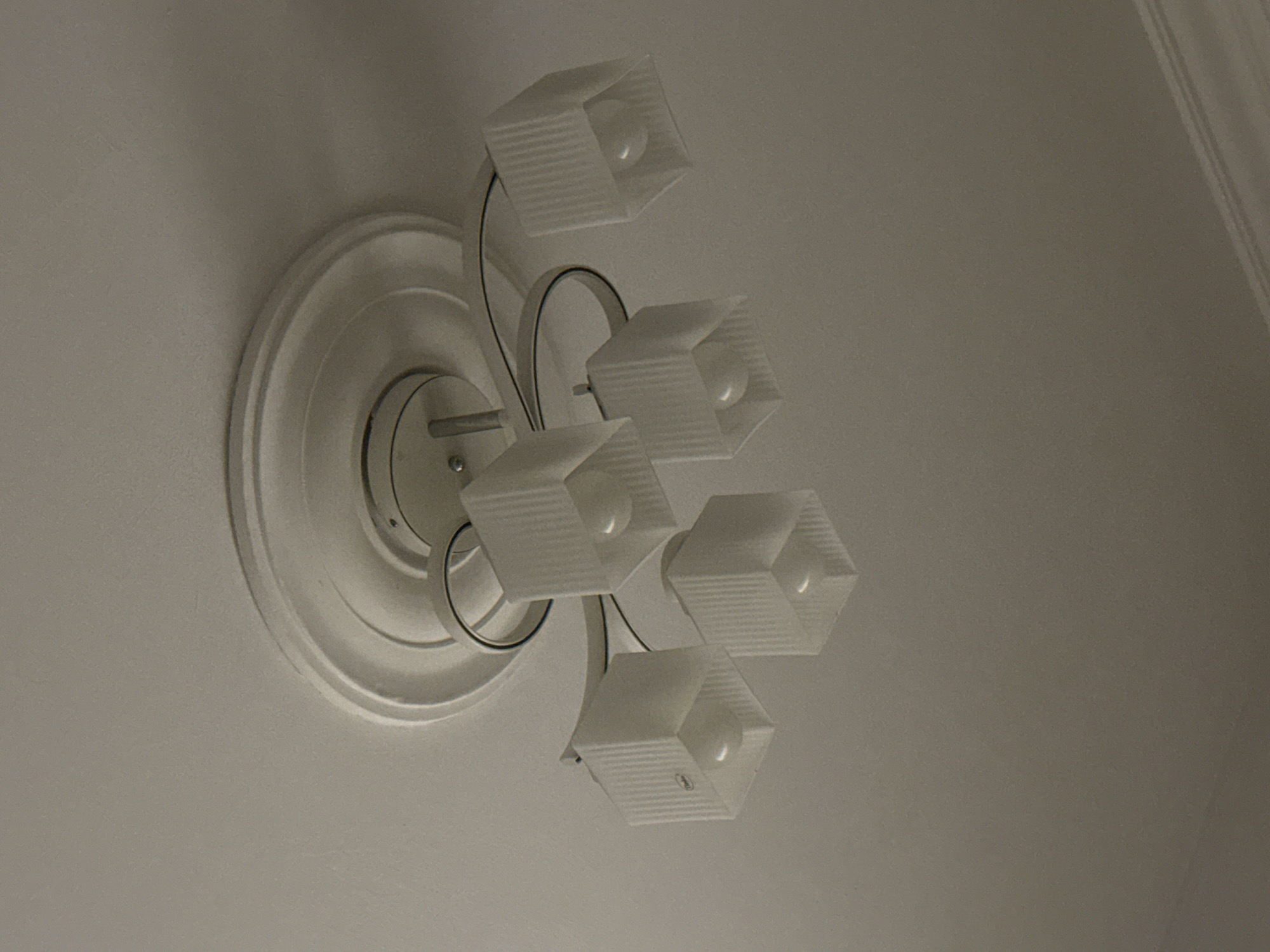This media file shows a close-up, slightly tilted-down view of a ceiling light fixture. The fixture has a decorative, circular base mounted on a white ceiling. Attached to the base are several arms that hold individual lamps. Each lamp is enclosed in a frosted, hexagonal-shaped shade with a ribbed texture. Inside each shade, a light bulb is visible. The overall scene suggests an indoor setting, likely a room in a residential or commercial building. The lighting appears to be artificial, as the light bulbs in the fixture are illuminated. There are no people visible in the media file. The focus is entirely on the lighting fixture itself and the surrounding ceiling. The angle of the shot gives a sense of looking up at the fixture. The colors are muted, primarily white and off-white from the fixture and ceiling, with some darker tones from shadows and the metallic parts of the fixture. The ribbed texture of the lampshades is a notable detail, creating a pattern of light and shadow.