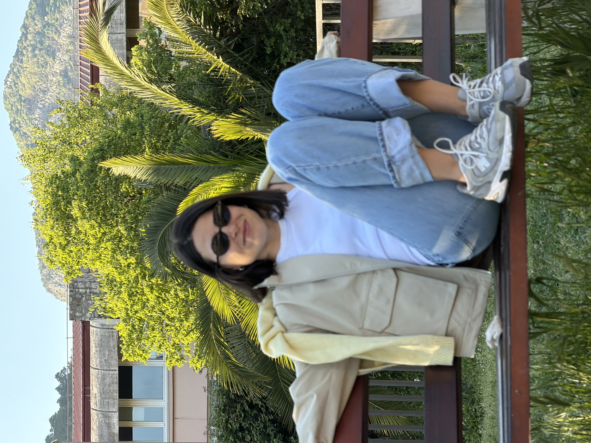 A person is sitting on a park bench in what appears to be Budva, Montenegro. The person, wearing a white t-shirt, jeans, sneakers, sunglasses, and a jacket, is reclined on the bench, with their legs elevated. The background includes trees and greenery, part of a building, and a mountain in the distance. The photo has been rotated, giving the appearance of the image being on its side.