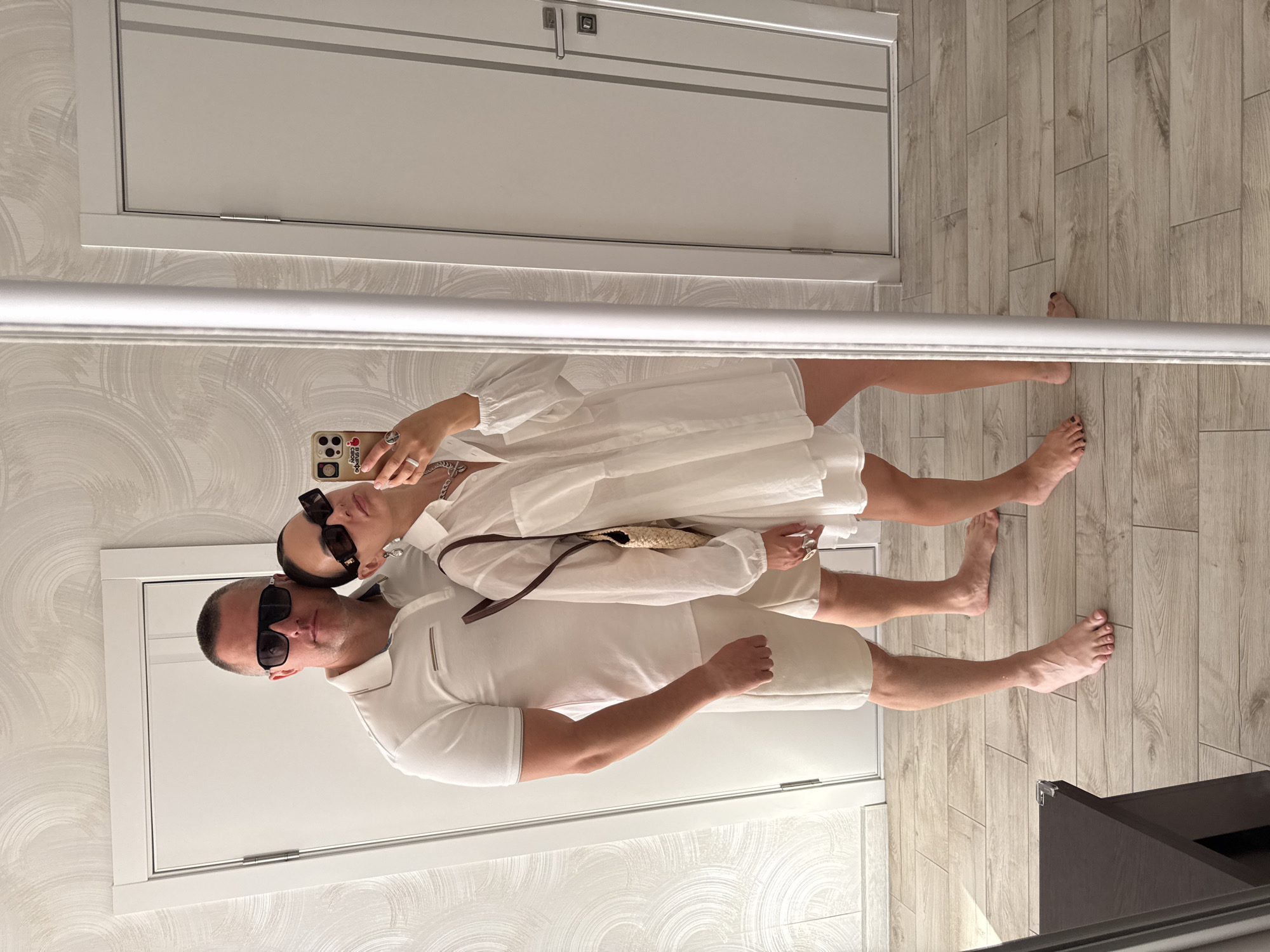 A couple is captured in a mirror selfie in a well-lit interior, likely a modern apartment or hotel room, with light-colored wood-plank flooring. The subjects are a man and a woman, both dressed in white, creating a cohesive aesthetic. The man, wearing sunglasses, is in the foreground, dressed in a white polo shirt and light-colored shorts. The woman, also wearing sunglasses and a white dress, stands slightly behind him, holding a smartphone to take the picture. Her outfit is complemented by a straw bag with a brown strap.
The room features white walls with a subtle textured pattern and two white doors. A prominent white molding runs horizontally across the image, dividing the upper and lower portions. The lighting suggests it's daytime, with no indication of weather. The overall mood is relaxed and summery.
There is no visible text in the image, except for what might be on the smartphone screen, which is too small to read. The reflection in the mirror indicates the presence of additional furniture, possibly a dark cabinet in the lower right corner. The couple appears content, with their poses suggesting a casual and intimate moment. The location context is Sochi, Russia.