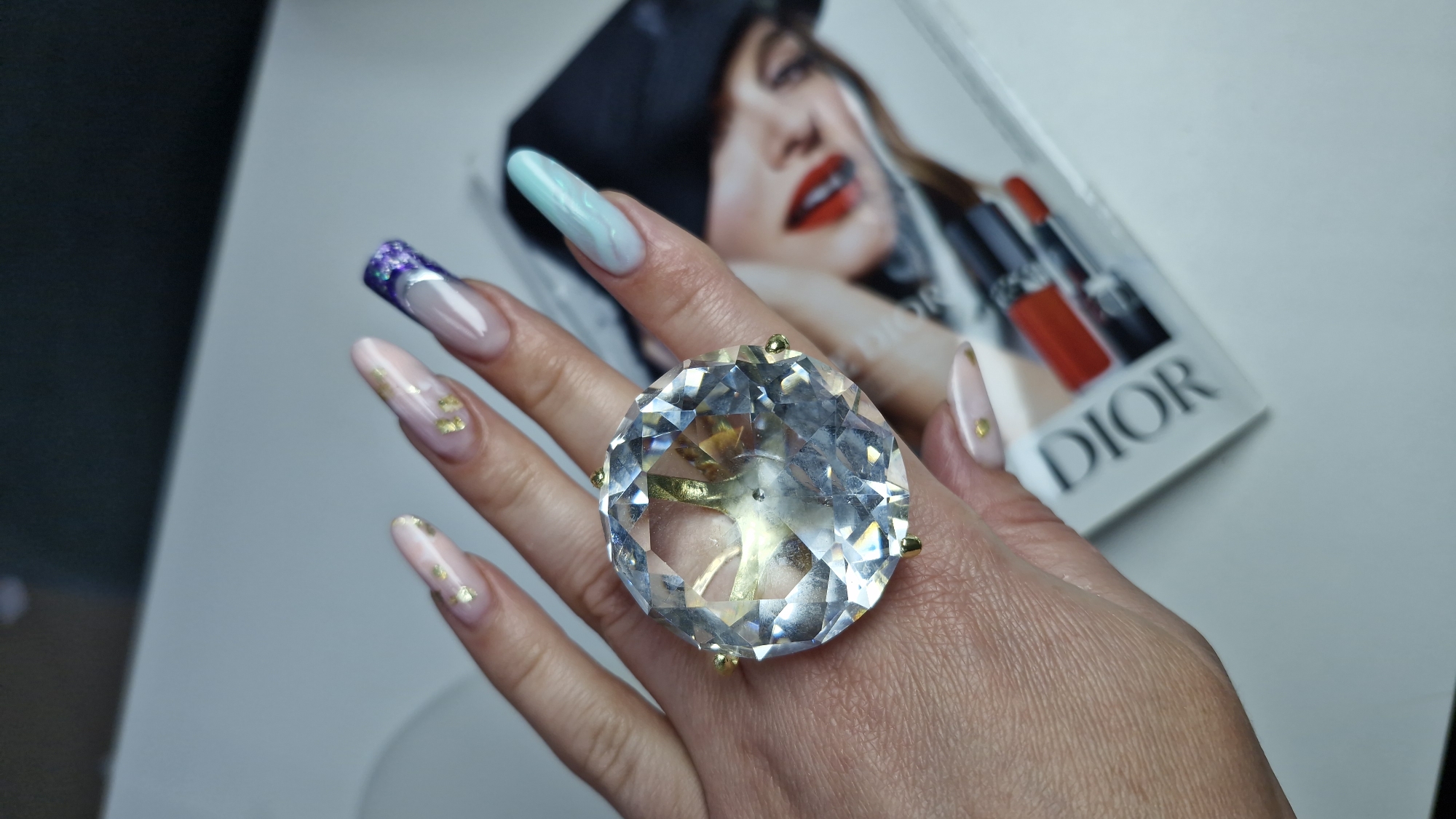 A woman's hand is prominently displayed, adorned with a large, clear, faceted crystal ring featuring gold-toned prongs. The fingernails are long and almond-shaped, with distinct designs: one has a purple glitter tip, another is light blue, and two have a pale pink base with gold foil accents.
The background includes a magazine, with a model wearing red lipstick visible in the periphery. The word "DIOR" is also discernible on the magazine.
The location of the scene is unknown and no evidence is included in the image that place it in Otchor, Russia.