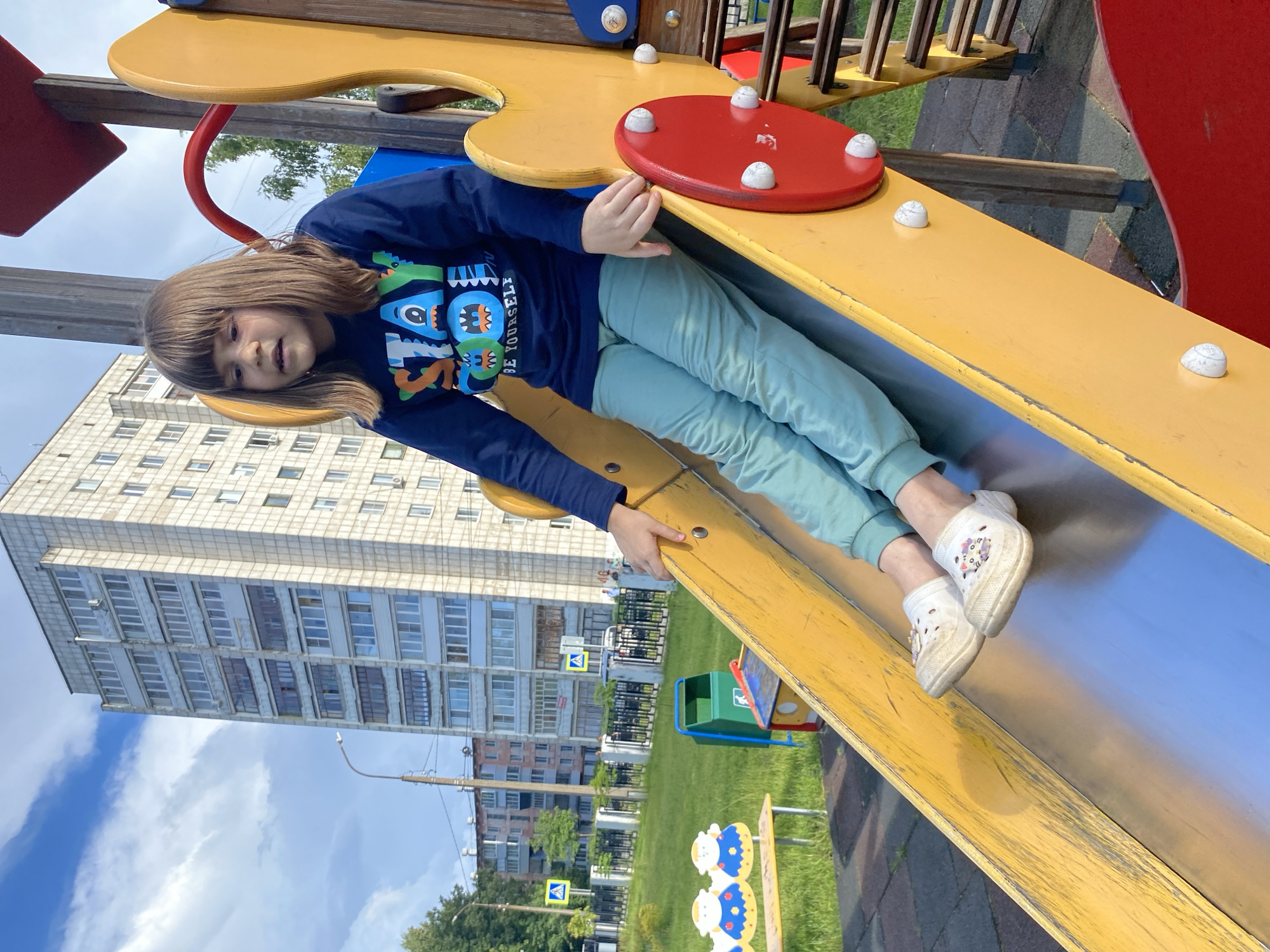 The image shows a young person lying on a yellow playground slide. The person is wearing a dark blue long-sleeved shirt with a graphic print of monsters and the text "BE YOURSELF" and light green pants. They are wearing white shoes with colorful designs on them. The slide is part of a larger playground structure with red and yellow elements. In the background, a tall, light-colored apartment building with many windows is visible against a blue sky with white clouds. There are trees and a grassy area with playground equipment in front of the building. The scene appears to be outdoors on a sunny day. The person's expression is somewhat neutral, with a slight smile. The time of day is likely daytime, given the bright lighting and blue sky. The image is oriented vertically, with the top of the frame on the left. The location context is Chaikovsky, Russia.