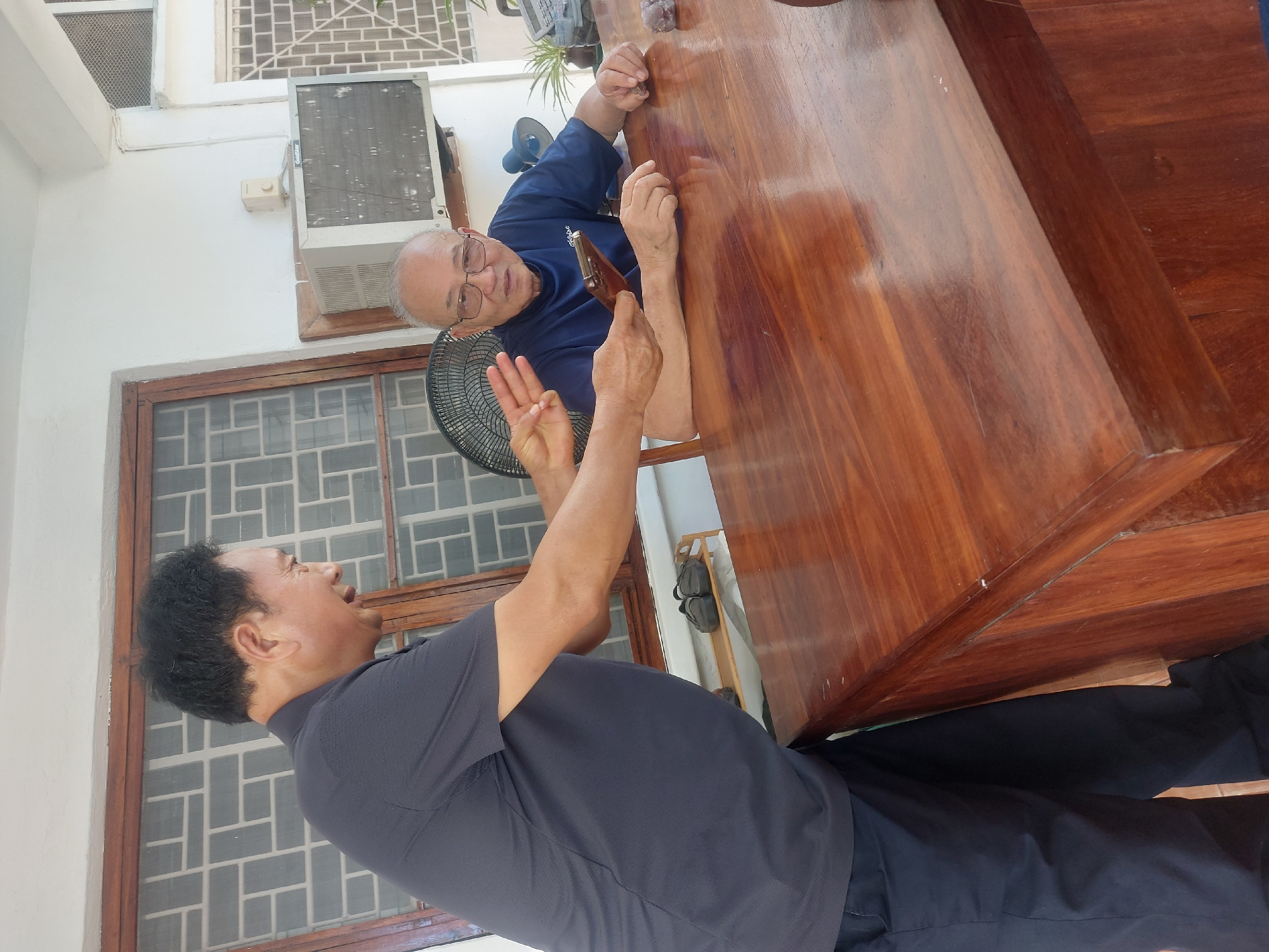 The image captures two adult men engaged in an animated conversation in what appears to be a casual indoor or semi-outdoor setting. The scene is brightly lit, suggesting it is daytime.
One man, older with light skin, balding with gray hair on the sides, and wearing glasses, is seated at a large, polished wooden table. He is dressed in a blue short-sleeved polo shirt, which features a "Goldstar" logo on the left chest. He leans forward on the table, looking attentively towards the other man. A small, dark, irregularly shaped object, possibly a stone or carving, rests on the table near his right hand.
Standing opposite him across the table is a middle-aged man with darker skin and dark hair, wearing a dark gray short-sleeved polo shirt and dark pants. His mouth is open, indicating he is speaking or laughing with enthusiasm. His left hand is raised, clearly displaying three fingers in a gesture, while his right hand holds a small, brown, elongated object, possibly a decorative item or tool, above the table.
The background features white walls, a large wooden-framed window with security grilles, and a black circular standing fan. An air conditioning unit, also bearing the "Goldstar" brand name, is mounted high on the wall. The prominent wooden table has a rich, reddish-brown hue and a reflective, polished surface. The presence of cooling appliances suggests a warm climate. The overall mood appears to be one of engaged and friendly interaction.