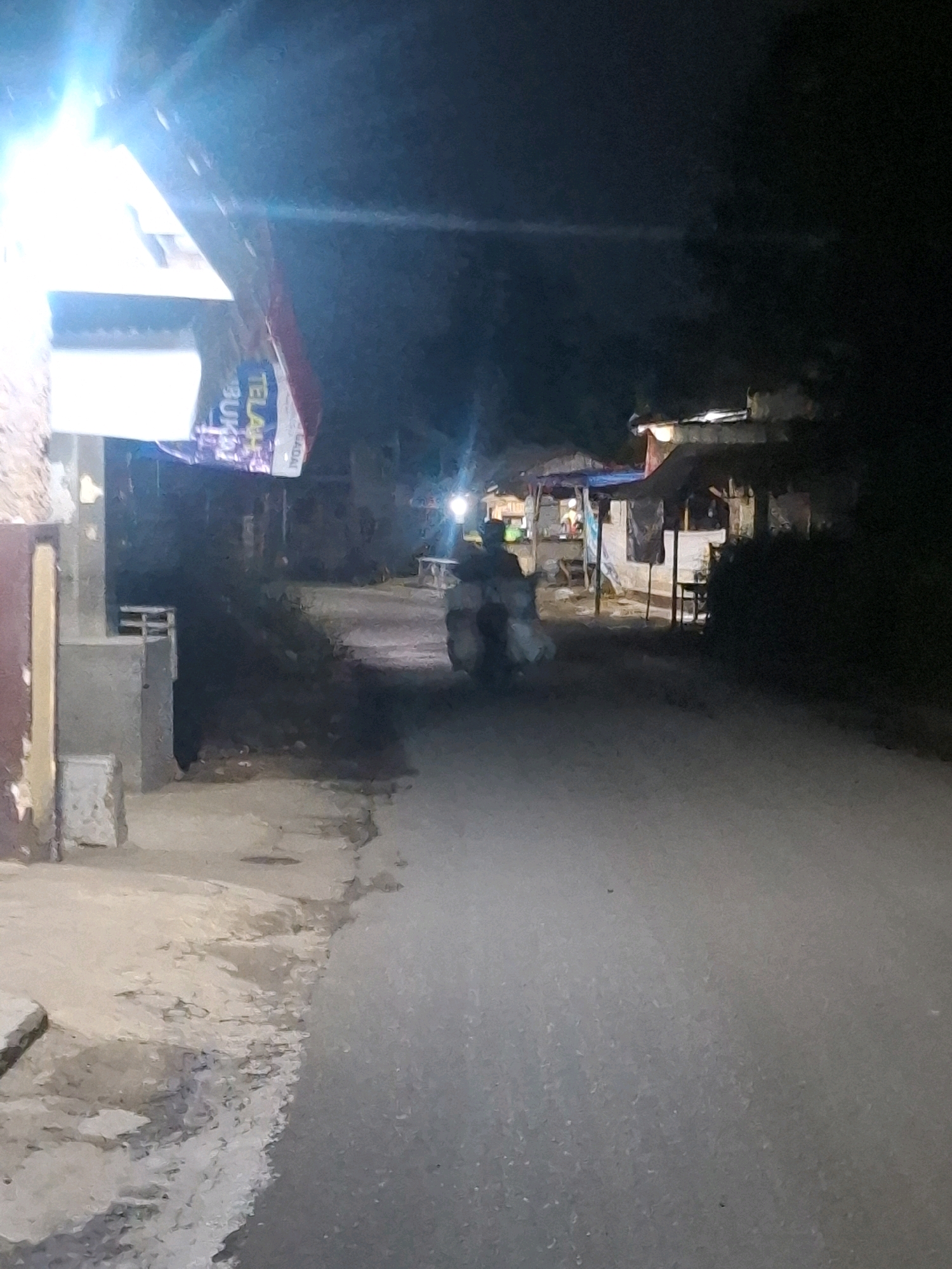 The image depicts a dimly lit street scene at night in Ciheulang Tonggoh, Indonesia. A motorcycle is visible in the middle of the road, with a rider and a large load of items strapped to the back. The motorcycle is illuminated by the headlights of an unseen vehicle, creating a bright, blown-out effect in the upper left portion of the image.
On the left side of the road, there is a building with a sign that reads "TELAH BUKA" which translates to "NOW OPEN" in Indonesian. The building appears to be a shop or a small business.
Further down the street, on the right side, are several small stalls or shops, also illuminated by lights, suggesting a marketplace or a commercial area. The overall impression is one of a bustling night scene in a local Indonesian community.
