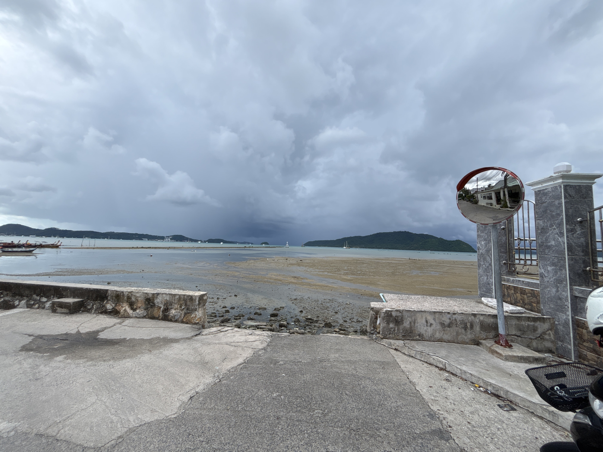 The image shows a view of the sea from a concrete embankment in Rawai, Thailand. The sky is overcast with dark clouds. The tide is low, revealing a muddy shoreline with scattered rocks and debris. In the distance, several boats are anchored on the water, and a lighthouse is visible on the horizon. To the right of the image, a convex mirror is mounted on a pole, reflecting a street scene with buildings. A part of a motorcycle with a black basket is parked in the foreground. The concrete embankment is cracked and weathered.