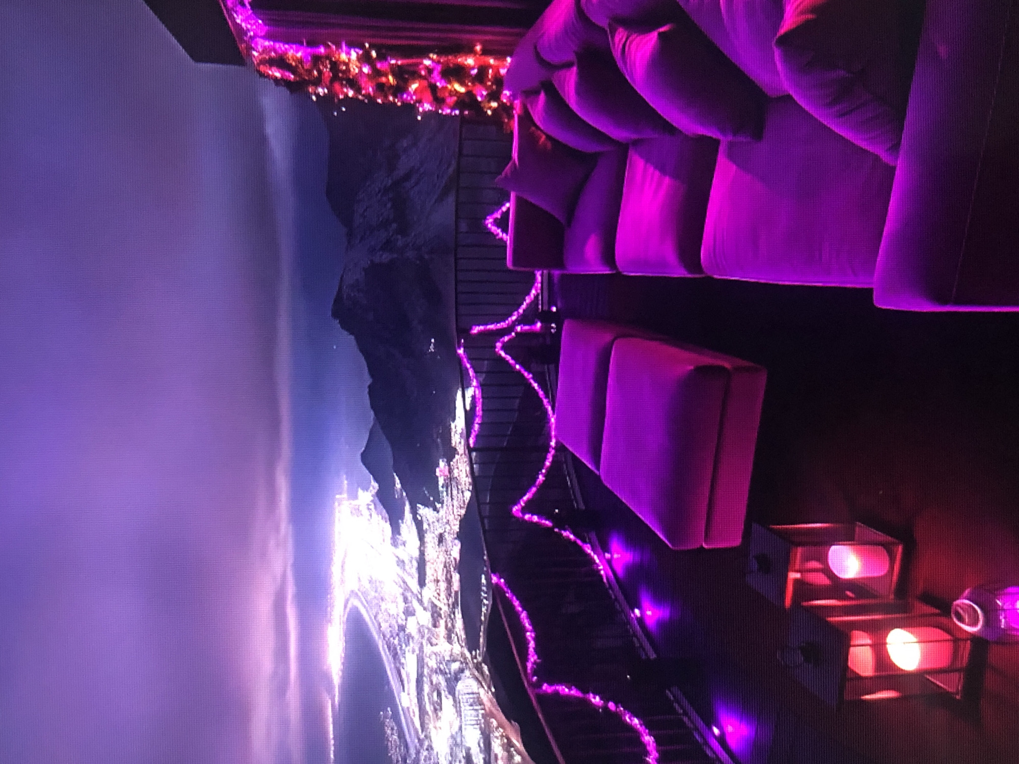 Here is a description of the image:
The image shows a rooftop lounge area with a panoramic view of what appears to be the city of Lhokseumawe, Indonesia, at night. The lounge is decorated with purple lights, including string lights along the railing. There is a large, plush sofa with many cushions and a smaller, individual seat. Some boxy light fixtures provide additional illumination, casting a reddish glow. The backdrop shows a city skyline with lights stretching along the coastline, next to a mountain. The sky is overcast.