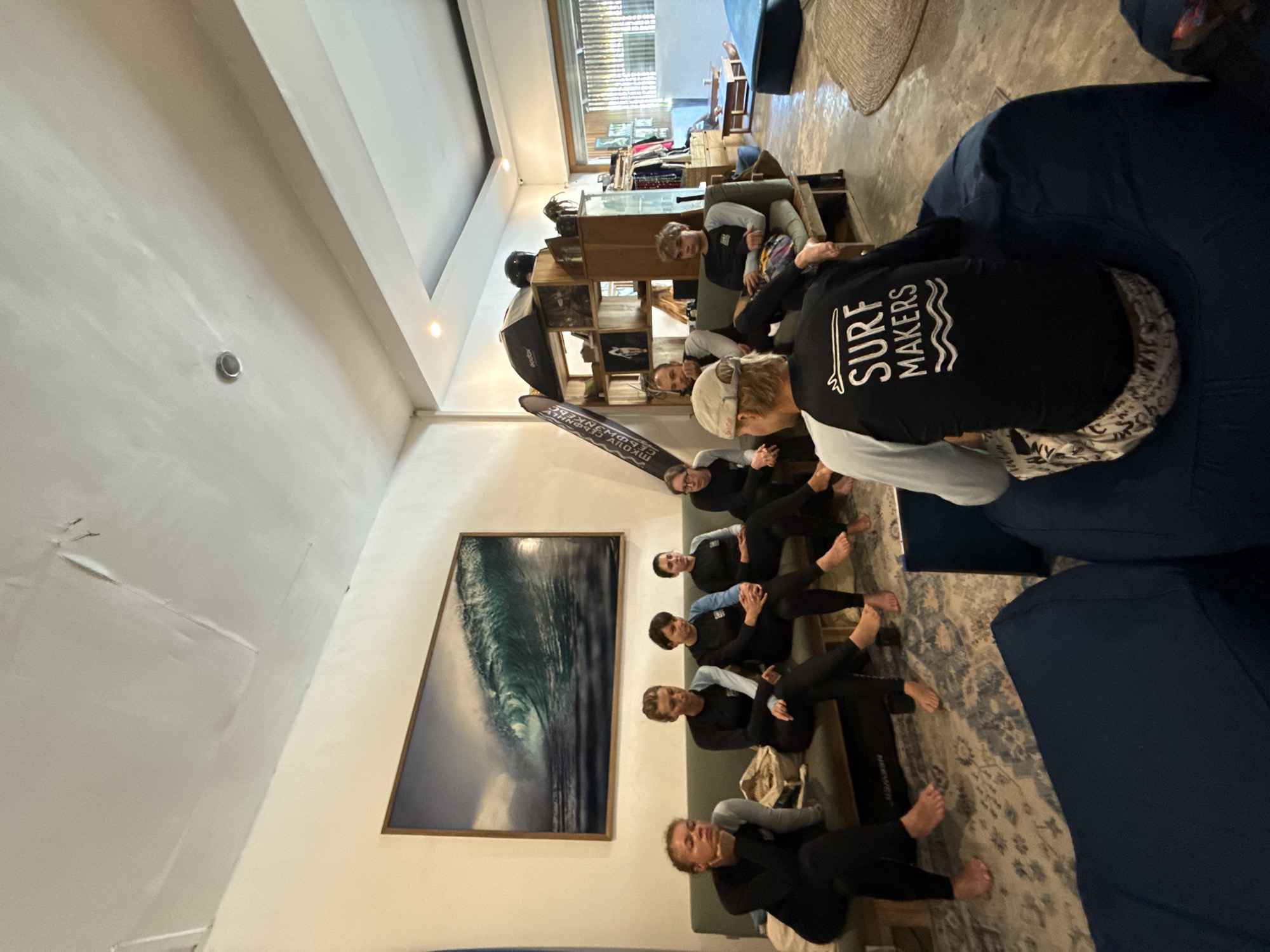 Inside a room, a group of people are seated in a row and on a couch. A large wave painting hangs on the wall. One person wears a shirt that says "Surf Makers." The location is likely Kuta, Indonesia, given the surfing theme and the style of the building.