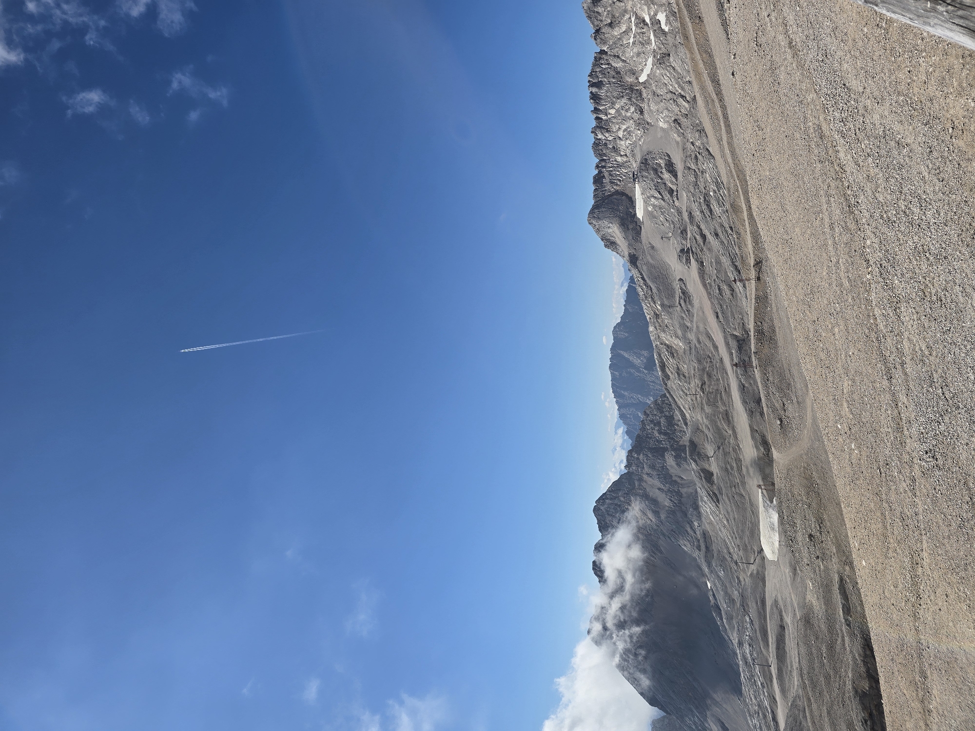 The image captures a high-altitude, mountainous landscape under a clear blue sky with a few wisps of clouds. A prominent contrail from an airplane stretches across the sky, indicating a flight path. The foreground is a barren, rocky slope with a light brown, gravelly texture. In the midground, there are more rocky mountain ranges, some with patches of snow or ice. Several ski lifts or cable car towers are visible, suggesting a recreational area, possibly for winter sports or summer hiking. A large, white rectangular structure, possibly a snow-making machine or a shelter, is situated on one of the slopes. The background features more rugged, gray mountain peaks, with some shrouded in light clouds. The overall impression is one of a vast, natural, and somewhat stark environment. This scene is likely the Zugspitze area, Germany's highest peak, located near Garmisch-Partenkirchen, known for its glaciers and high-altitude tourism. There are no people visible in the image.