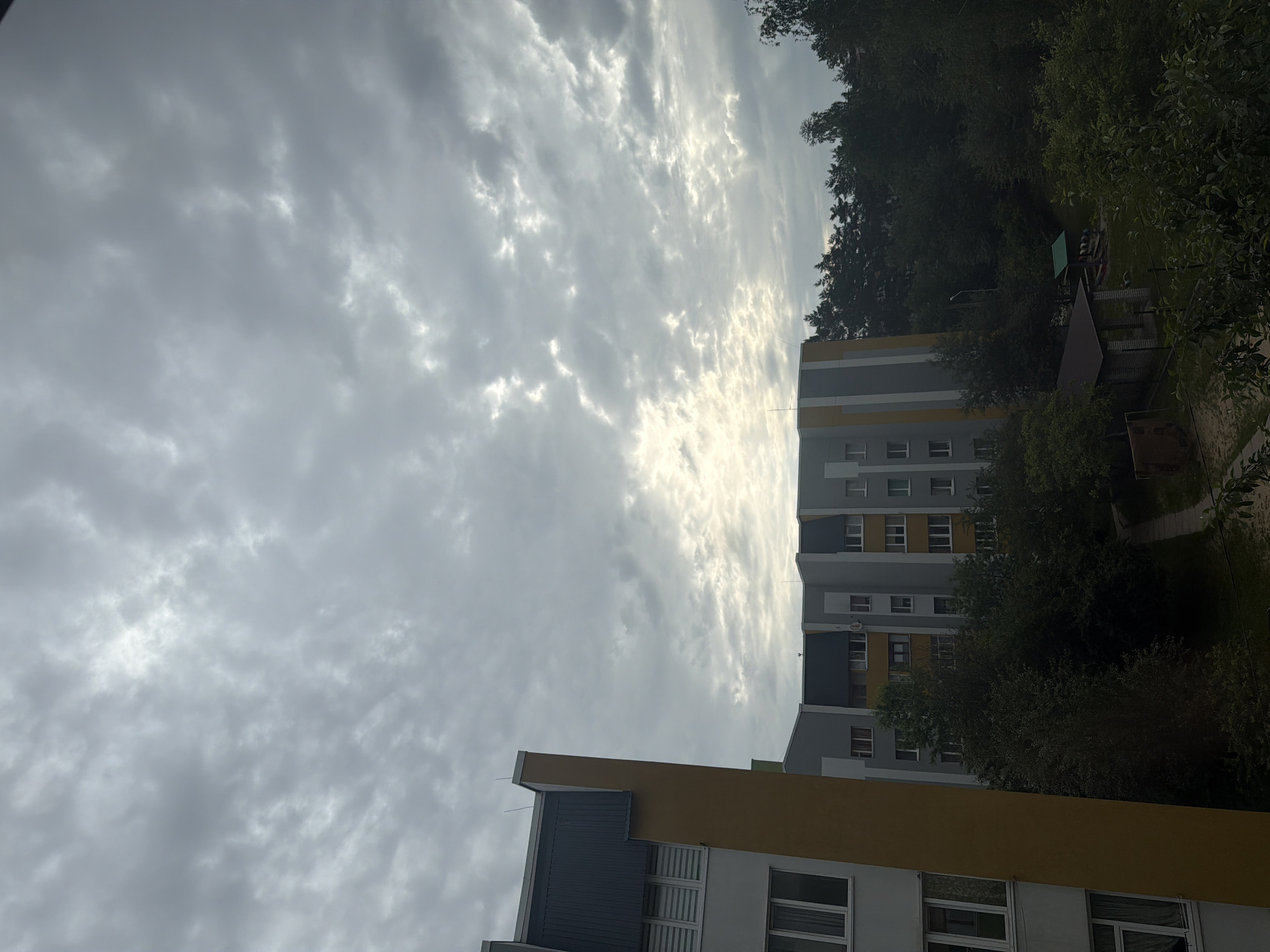This is a daytime, slightly overcast outdoor scene captured from an elevated perspective, likely a window, overlooking a residential area in Novoiavorivsk, Ukraine. In the foreground, to the left, is the side of a multi-story building with a yellow and grey facade and a dark blue sloped roof. Its windows are visible, some with blinds. To the right of this building, further in the background, is another multi-story building with a grey and yellow facade and a dark blue sloped roof. Trees and greenery dominate the middle ground, obscuring parts of the lower levels of the buildings. A small playground with a sandpit and a shelter can be seen amongst the trees. The sky above is filled with a textured layer of grey clouds, with patches of brighter light indicating the sun's presence filtering through. The overall impression is of a calm, everyday urban environment under a cloudy sky. There are no people or animals visible in the image.
