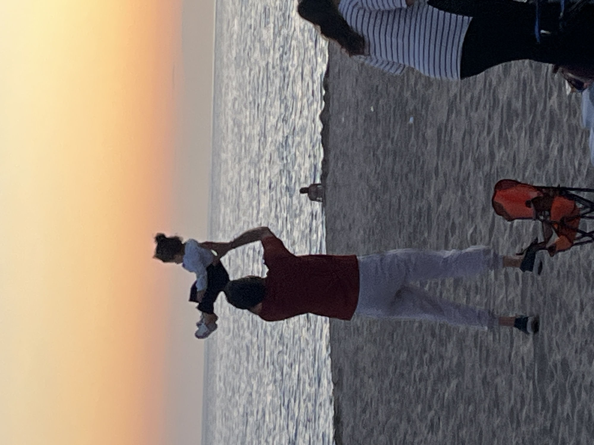 The image depicts a beach scene at sunset. A man in a red shirt and light-colored pants is holding a young child up in the air. The child is wearing a light-colored shirt and dark pants. Behind them is the ocean, reflecting the colors of the sunset. To the right, on the sandy beach, is a person in a striped shirt and pants and a red beach chair. Another person is in the background. The location is likely Alanya, Turkey, based on the light and surrounding area.