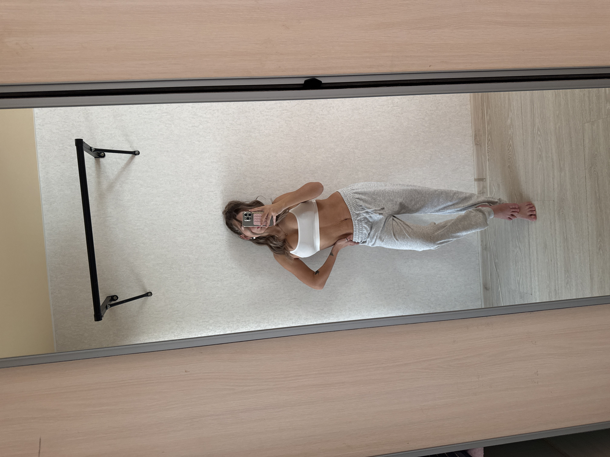 An indoor scene is captured as a mirror selfie. The specific location within Chaikovsky, Russia, is unidentifiable from the image.
**Subject:** A female is depicted from mid-torso to feet, reflected in a large rectangular mirror. Her face is partially obscured by a smartphone, held in her raised left hand, with which she is taking the photograph. She has light-colored, wavy hair, wears earrings and a necklace, and a tattoo is visible on her left forearm. Her right hand rests on her hip.
**Attire:** She is wearing a white crop top and grey sweatpants with a visible drawstring. She is barefoot.
**Environment (reflected):** The background consists of a light-colored, textured wallpapered wall. A black horizontal exercise bar with two diagonal supports is mounted on this wall, positioned above the subject's head. The floor is composed of light wood laminate. A beige ceiling is visible at the top of the reflection.
**Objects:** The primary object is the large rectangular mirror, which is framed by a light wood-grained panel on the left and a dark vertical trim on the right, consistent with a wardrobe door or a large freestanding mirror. The female is holding a smartphone, which has a light-colored, patterned case.