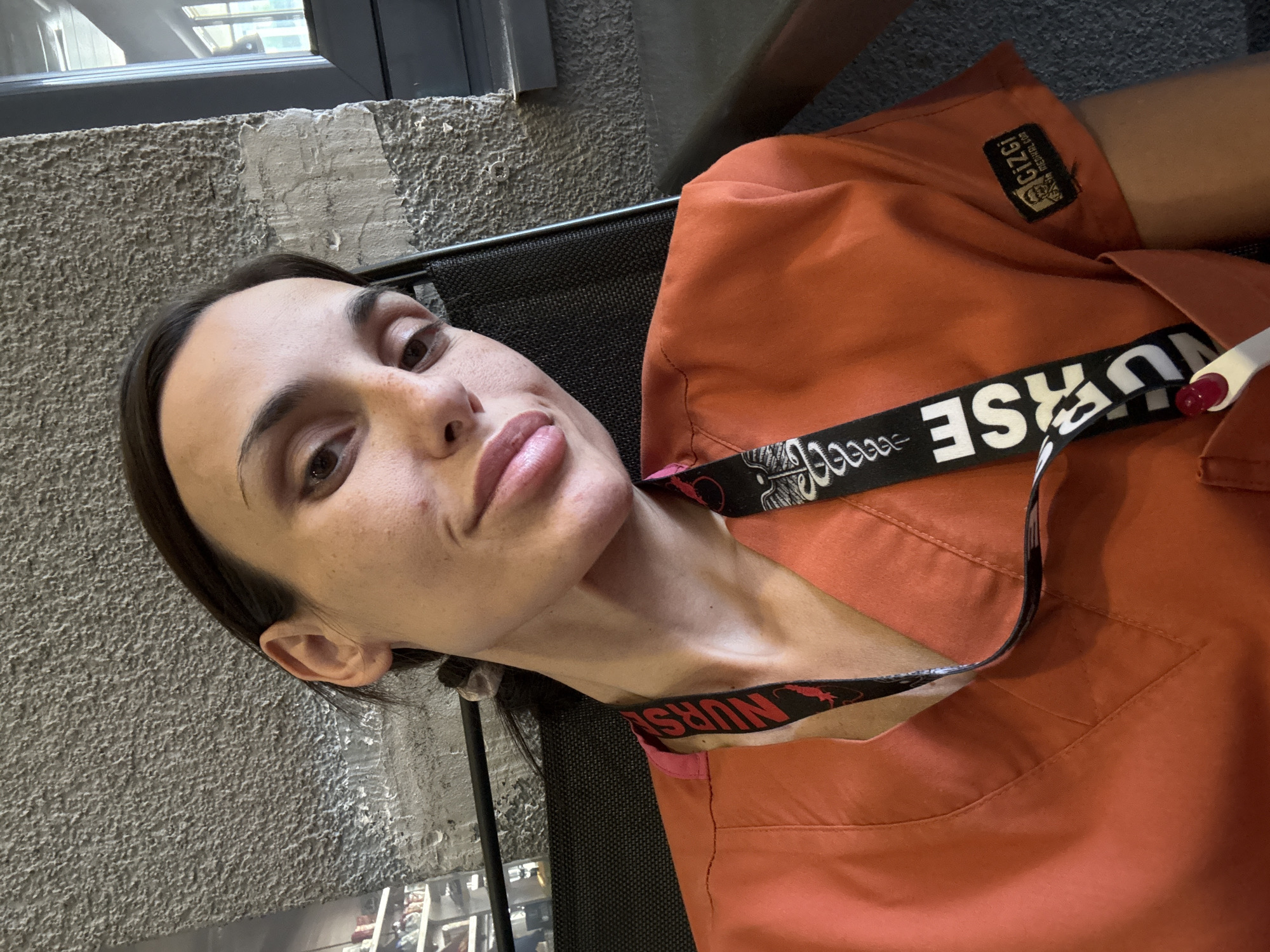 A young woman, likely a nurse, is captured in a close-up, waist-up photograph. She is seated, with her head turned towards the camera, offering a slight smile and a direct gaze. She is wearing bright orange scrubs, a common attire for medical professionals, and a lanyard with "NURSE" printed in large white letters around her neck. The background is a textured, gray concrete wall. To the right, a large window frames a view of the outdoors, suggesting an urban setting. The lighting is natural, casting soft shadows. The woman's facial features are clear, and her makeup is subtle. The overall impression is one of relaxed professionalism. The image appears to be taken in Pleven, Bulgaria.