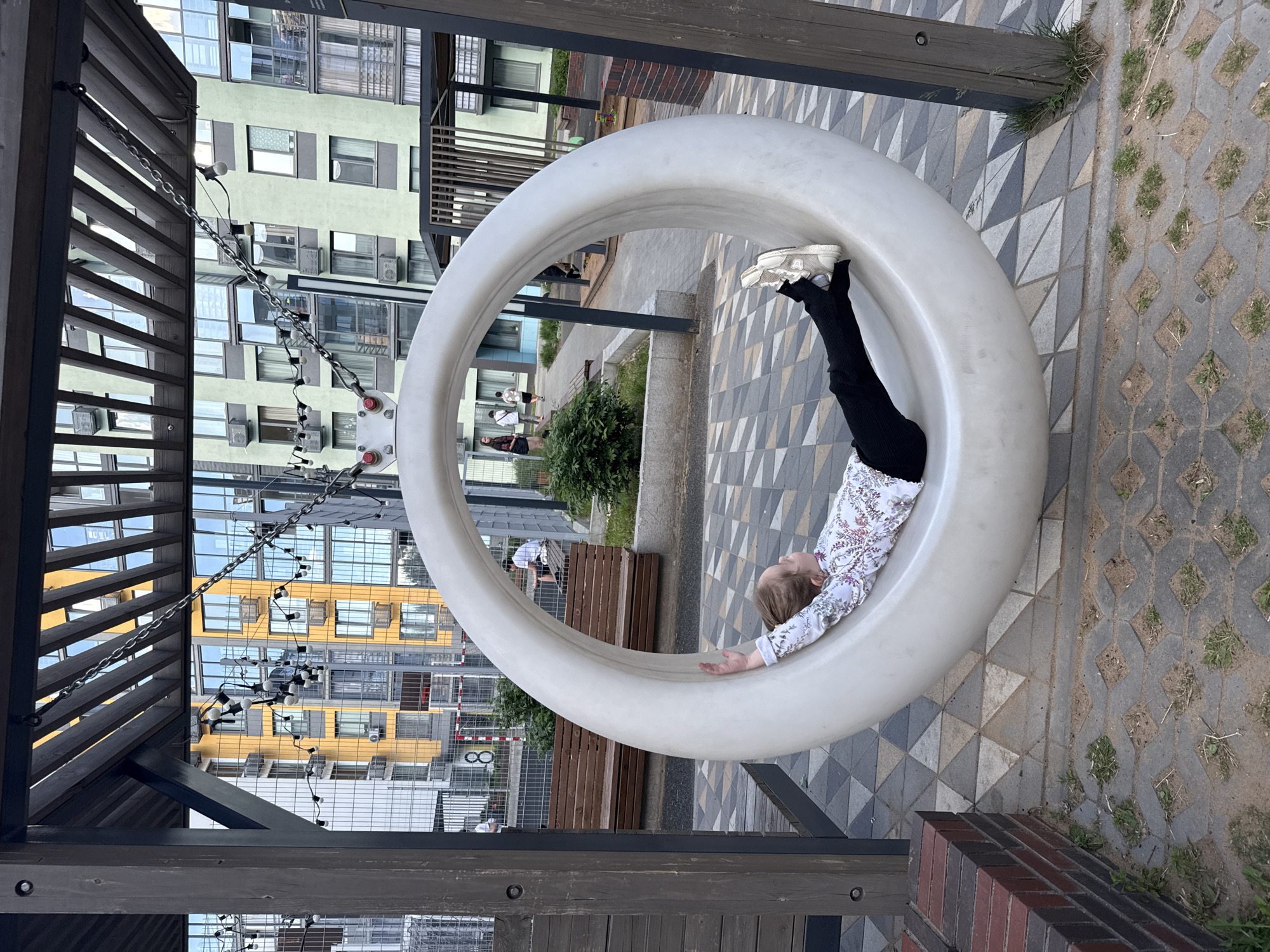 The image displays an outdoor scene within a modern residential complex courtyard, located in Izhevsk, Russia. The primary subject is a young girl, positioned lying on her back within a large, smooth, white circular swing. The swing is suspended by metal chains from a dark wooden and metal pergola structure overhead, featuring wooden slats and sturdy posts. The girl wears a long-sleeved white shirt with a multi-colored floral or leaf pattern, black leggings or pants that cover her feet, and light-colored athletic shoes. Her left arm is slightly raised and extended, resting on the inner edge of the swing. Her feet are propped against the opposite edge.
In the background, multiple multi-story apartment buildings are visible; one on the left is bright yellow with numerous windows and balconies, while another on the right is pale green, also featuring external air conditioning units. String lights with small bulbs are draped across the pergola structure and along some chains. The ground is predominantly paved with interlocking geometric tiles in shades of light and dark grey. Towards the bottom of the frame, concrete pavers with patches of grass and exposed earth are present. A low wall constructed from red and dark grey bricks is visible in the lower left foreground.
Several other individuals are present in the mid-ground: one person is seated on a wooden bench behind the swing, and multiple people are walking along a paved pathway further back near a metal mesh fence. A white post with red markings, possibly indicating a sports area, is visible behind the fence. Small green bushes are scattered throughout the courtyard, and a sandpit is partially visible in the background, to the right of the girl.
