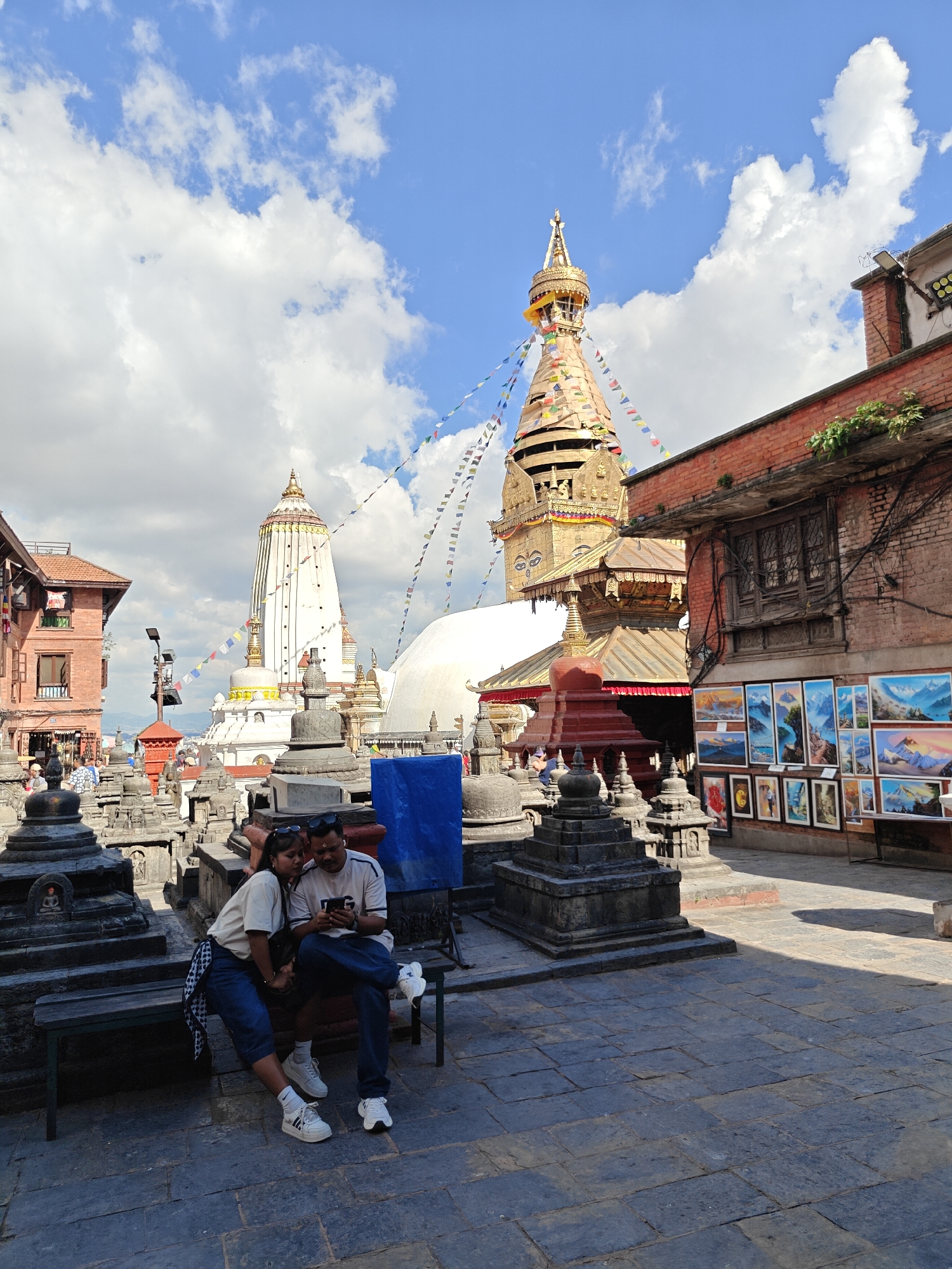 The image depicts a courtyard with several stupas, Buddhist prayer flags, and buildings, likely within the Swayambhunath Stupa complex in Kathmandu, Nepal. In the foreground, a young couple is seated on a bench, looking at a smartphone. The man is wearing a white t-shirt and blue jeans, while the woman is wearing a cream-colored t-shirt and blue jeans, with a checkered scarf around her waist. They are both wearing sneakers. Behind them, numerous grey stupas of varying sizes are arranged in rows. Further in the background, two prominent stupas are visible: a large white stupa with golden trim and a golden stupa topped with a spire and prayer flags. A brick building with windows and an attached display of paintings is to the right of the golden stupa. The sky is blue with scattered white clouds. The ground is paved with dark grey stones.