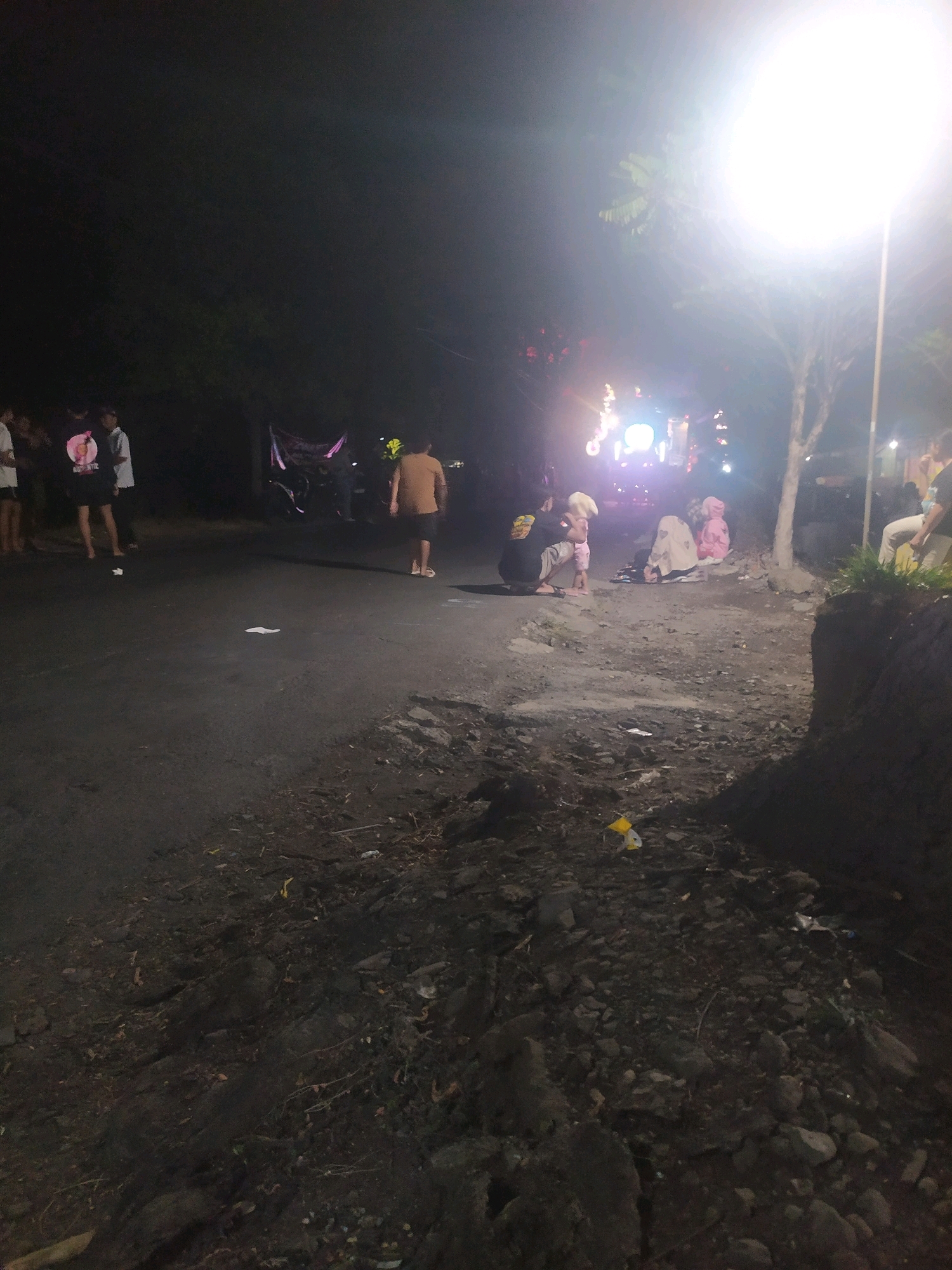The image depicts a nighttime scene on a paved road with a dirt shoulder, likely in Winong, Indonesia, given the provided context. A bright street lamp illuminates the right side of the frame, casting long shadows. Several individuals are present in the scene.
On the left side of the road, four people are standing, appearing to be observing something off-camera. One person wears a dark t-shirt with a graphic design and light-colored shorts. Another person to their left wears a light-colored t-shirt and shorts. The other two individuals on the far left are less clearly discernible.
Further down the road, a person in a light brown t-shirt and dark shorts is walking away from the camera towards the center of the road.
In the middle ground, a group of people are gathered near a brightly lit structure, possibly a stage or a vehicle with lights. A person in a dark shirt is kneeling and interacting with a young child. Two other individuals, one wearing a pink garment and the other a lighter colored garment, are seated or kneeling.
The environment appears to be somewhat unkempt, with debris, including rocks and what looks like scattered litter such as a yellow plastic bag, present on the dirt shoulder and along the edge of the road. A large tree is visible near the street lamp. The overall lighting is dim, with the exception of the intense street lamp and the lights from the structure in the middle ground.
