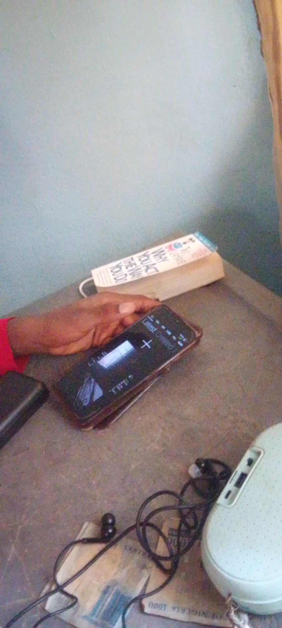 An overhead, medium shot shows a person's left hand resting on a table. The hand, with dark skin and short fingernails, is holding a black smartphone. The smartphone's screen displays text that reads "Latest Created" followed by some icons and a plus symbol. A portion of a book with the title "WHY YOU ACT THE WAY YOU DO" is visible behind the phone. To the left of the phone, a black rectangular object, possibly a power bank, is partially visible. In the lower right of the image, a light teal, oval-shaped object with perforated sides, resembling a retro telephone handset, is present, with a coiled black cord connecting to a black earpiece. The table surface appears to be made of a rough, mottled material. In the background, a light blue wall is visible.