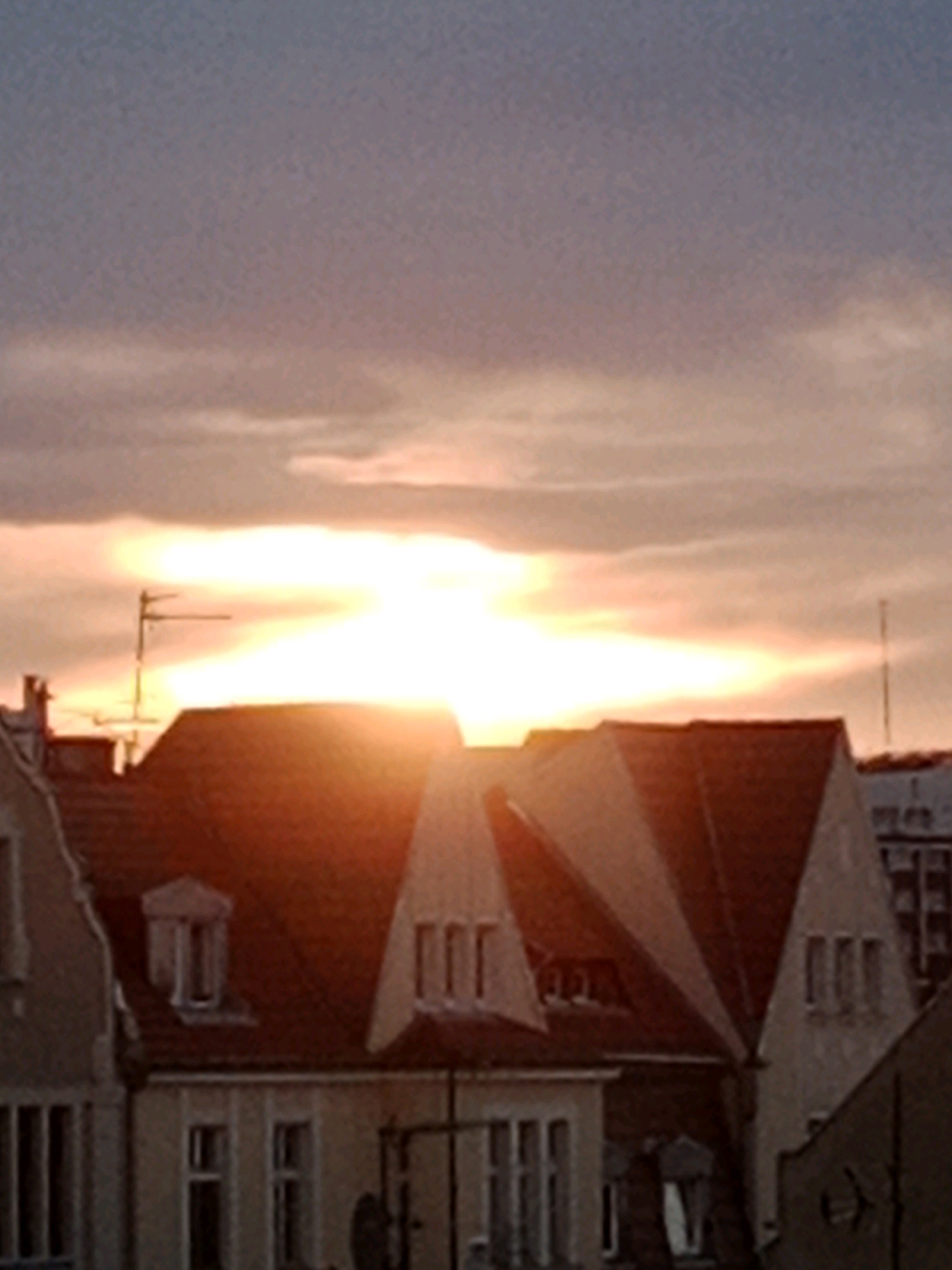 The image shows a sunset over a city skyline. The sky is filled with orange and pink clouds, and the sun is setting behind a row of buildings. The buildings appear to be old, with red tiled roofs and traditional architecture. In the foreground, there are several houses with visible windows and dormer windows. A television antenna is visible on top of one of the houses. The overall impression is one of a peaceful and picturesque evening in a European city. Based on the architectural style and the presence of a television antenna which suggests a residential area, the scene is consistent with an urban or suburban setting in Bydgoszcz, Poland.