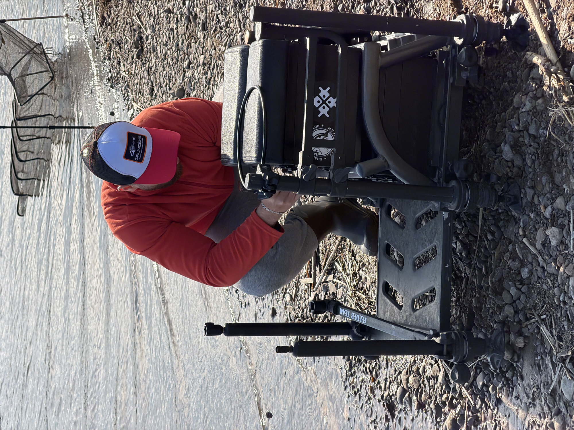 The image shows a man fishing by a river. The man is wearing a red long-sleeved shirt, grey pants, and a white, red, and black baseball cap. He is kneeling beside a black fishing seat with the words "FEEDER TEAM" on it. A fishing net is partially visible next to the river. The ground is covered in pebbles. It is impossible to determine if this scene is in Chaykovsky, Russia, from the image alone.