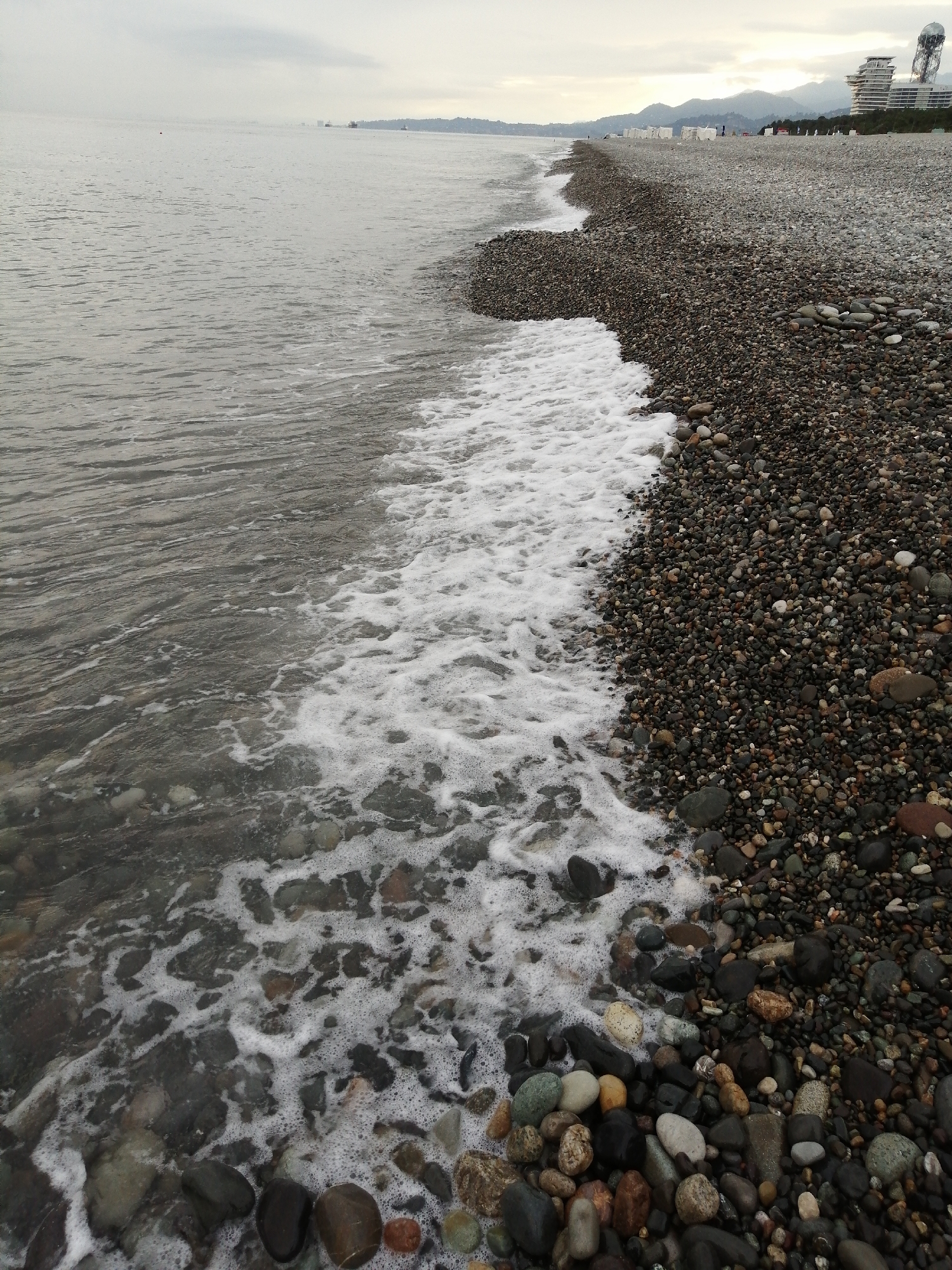This is a photograph of the pebble beach and the sea in Batumi, Georgia. The foreground shows the edge of the sea with waves lapping onto a shore covered in dark, multicolored pebbles of varying sizes. The water is a murky grey-green and the waves are creating white foam as they meet the shore. In the midground and background, the pebble beach extends further inland, and distant buildings and hills are visible under a cloudy sky. The overall impression is of a slightly overcast day at the seaside.