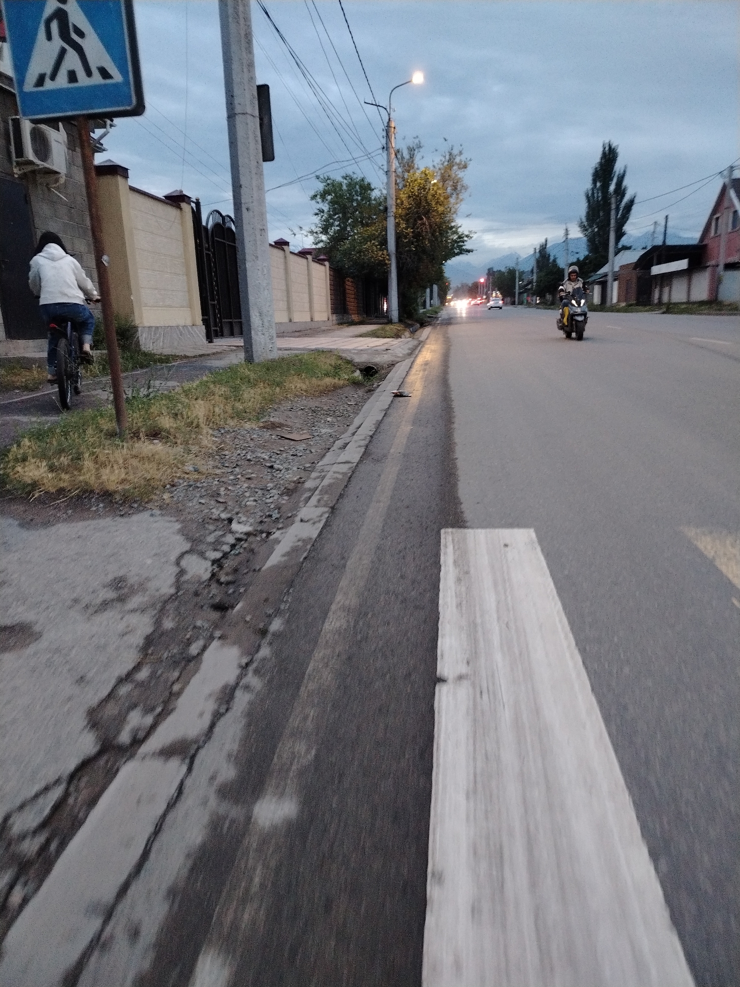 The image depicts a street scene at dusk in Bishkek, Kyrgyzstan. A cyclist in a white hoodie and blue jeans is riding a bicycle on the left side of the road, near a pedestrian crossing sign. A motorcycle with a rider wearing a helmet is traveling on the right side of the road. The street is paved, with some cracks and debris on the side. Buildings and trees line the street, and power lines are visible overhead. The sky is cloudy, and distant mountains can be seen. The lighting suggests it is either early morning or late evening.