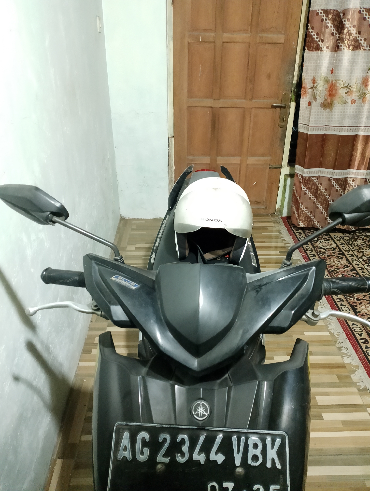 A white Honda helmet rests on the front of a black motorcycle. The motorcycle has a license plate that reads "AG 2344 VBK". The motorcycle is parked on a tiled floor next to a wooden door. A patterned curtain hangs on the right side of the door. The wall to the left of the motorcycle is painted white. This scene is located in the city of Rembang, Indonesia.