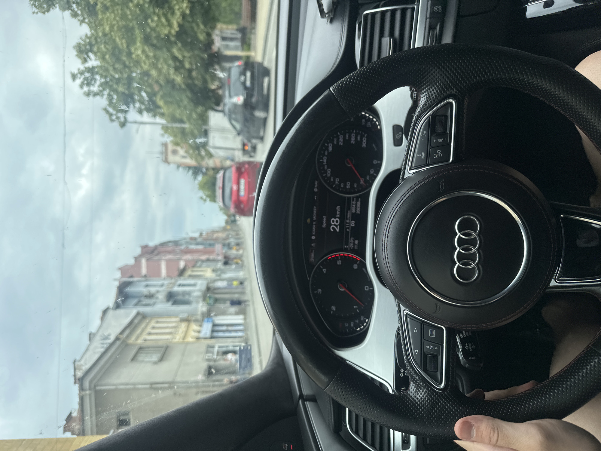 The image shows a first-person perspective from inside an Audi vehicle, looking forward through the windshield at a street scene in Kazanlak, Bulgaria. The driver's hands are visible on the steering wheel, and the dashboard displays the car's speed at 28 km/h. The street is lined with multi-story buildings, some with colorful facades. A red car is in front, followed by a black car and a truck. The sky is overcast. The environment appears to be an urban setting in Kazanlak, Bulgaria, with a moderate amount of traffic on a cloudy day.