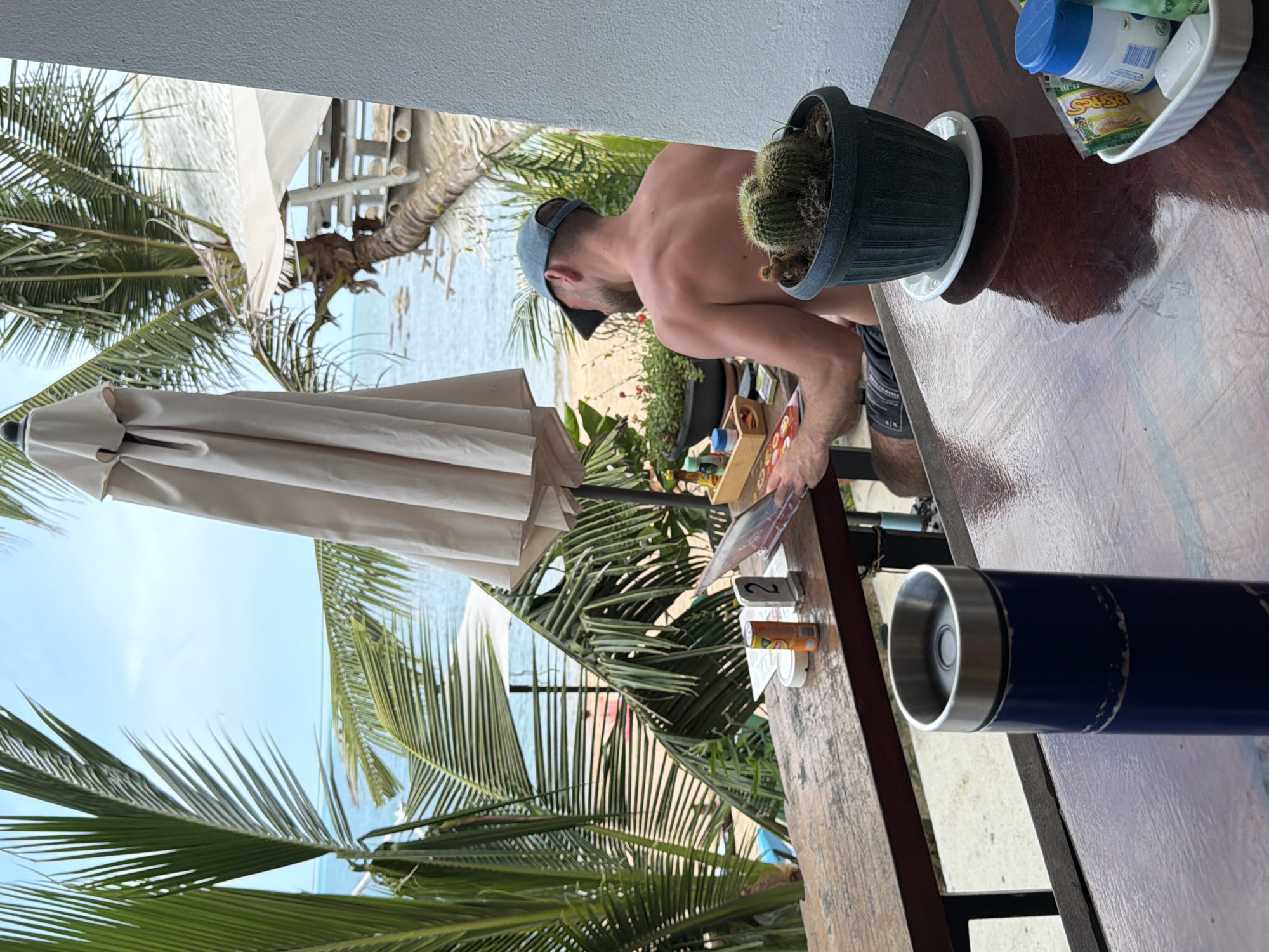A man wearing a blue baseball cap and dark shorts is seated at a wooden table, facing away from the camera. He is looking at a magazine or menu. In front of him on the table are a small orange can, a table number stand with a "2" on it, and a wooden box containing condiments. To the right of the man, a potted cactus sits on a small white saucer. The table is outdoors and appears to be on a balcony or patio overlooking a beach. Palm trees and a closed umbrella are visible in the background, along with the ocean and a clear sky. The scene is set in a tropical location, likely a restaurant or cafe near the beach on Ko Samui District, Thailand.