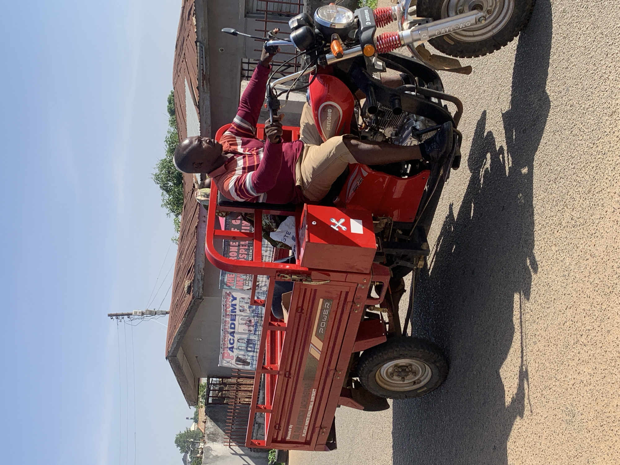 The image depicts a man sitting on a red tricycle, possibly a cargo tricycle, in a street setting. The man is wearing a maroon and white striped shirt and tan shorts. The tricycle has a "POWER" logo and the word "DAYLONG" on the front. Behind the tricycle, there is a building with a sign for "Precious Stone Academy". The scene appears to be in Jos, Nigeria, as suggested by the text "PLATEAU NORTH SENATORIAL DISTRICT" on the side of the tricycle. The sky is clear and sunny. The tricycle is on an asphalt road.
