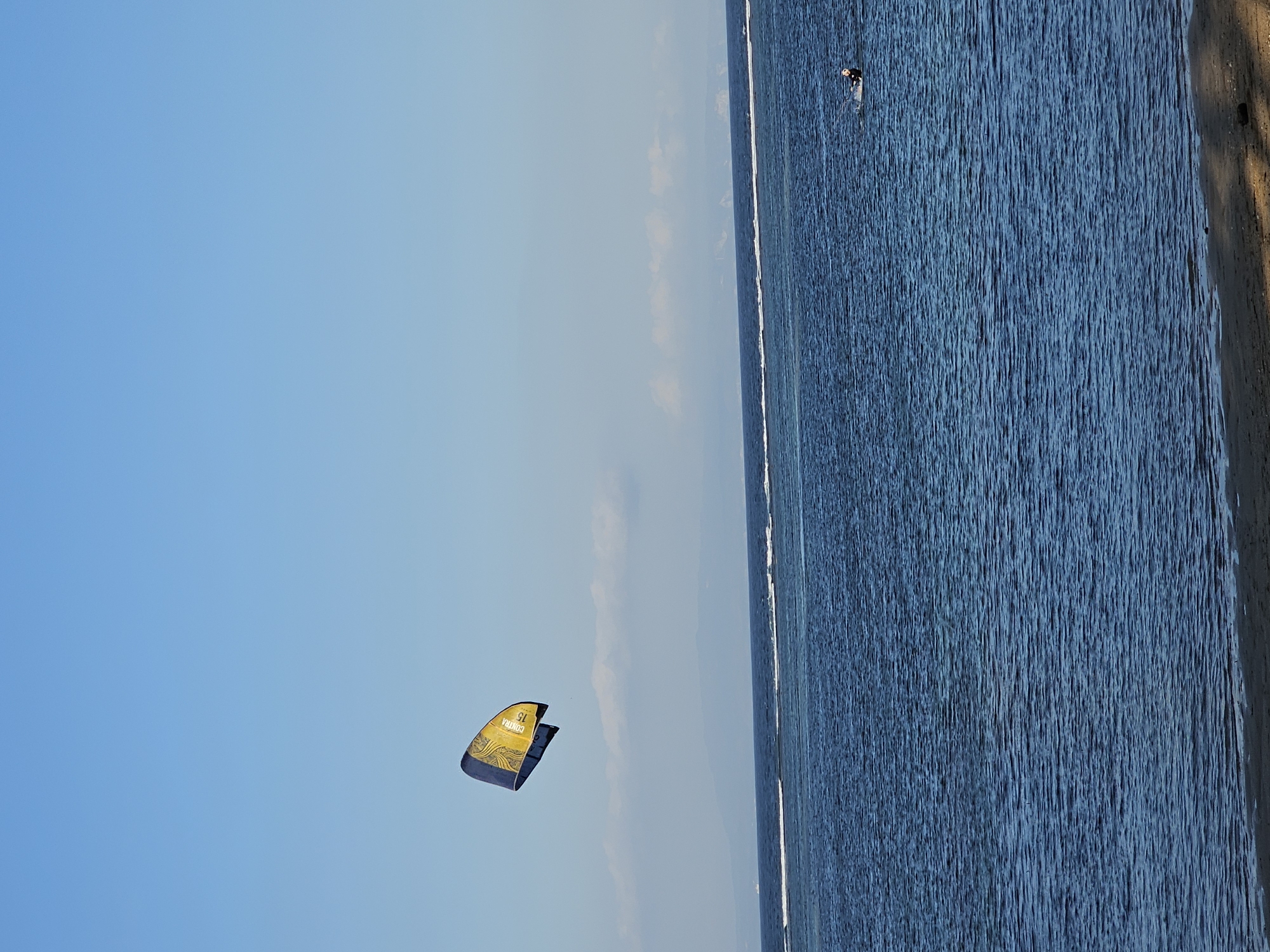 The image captures a serene seascape at the beach in Denpasar, Indonesia, under a clear blue sky. In the vast expanse of the ocean, a kiteboarder is engaged in their sport, with the kite, a distinct yellow and blue, soaring high in the air. The kiteboarder is visible in the water, in the right side of the frame, appearing to be in motion. Gentle waves create a textured surface on the deep blue water, contrasting with the thin white line of a wave break further out. In the distance, the hazy outline of land and clouds suggests a coastline. The foreground shows a sandy beach with a hint of shadows, indicating sunlight.
