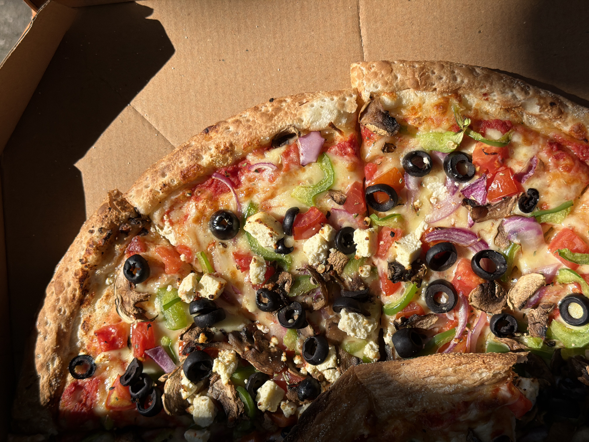 A close-up, overhead shot shows a pizza in a brown cardboard box. The pizza is cut into slices and topped with tomatoes, olives, mushrooms, green peppers, red onions, and feta cheese. The pizza crust is golden brown and has a slightly charred texture. The lighting suggests it is daytime, with sunlight casting shadows across the box and pizza. The location is the city of Mengwi, Indonesia.