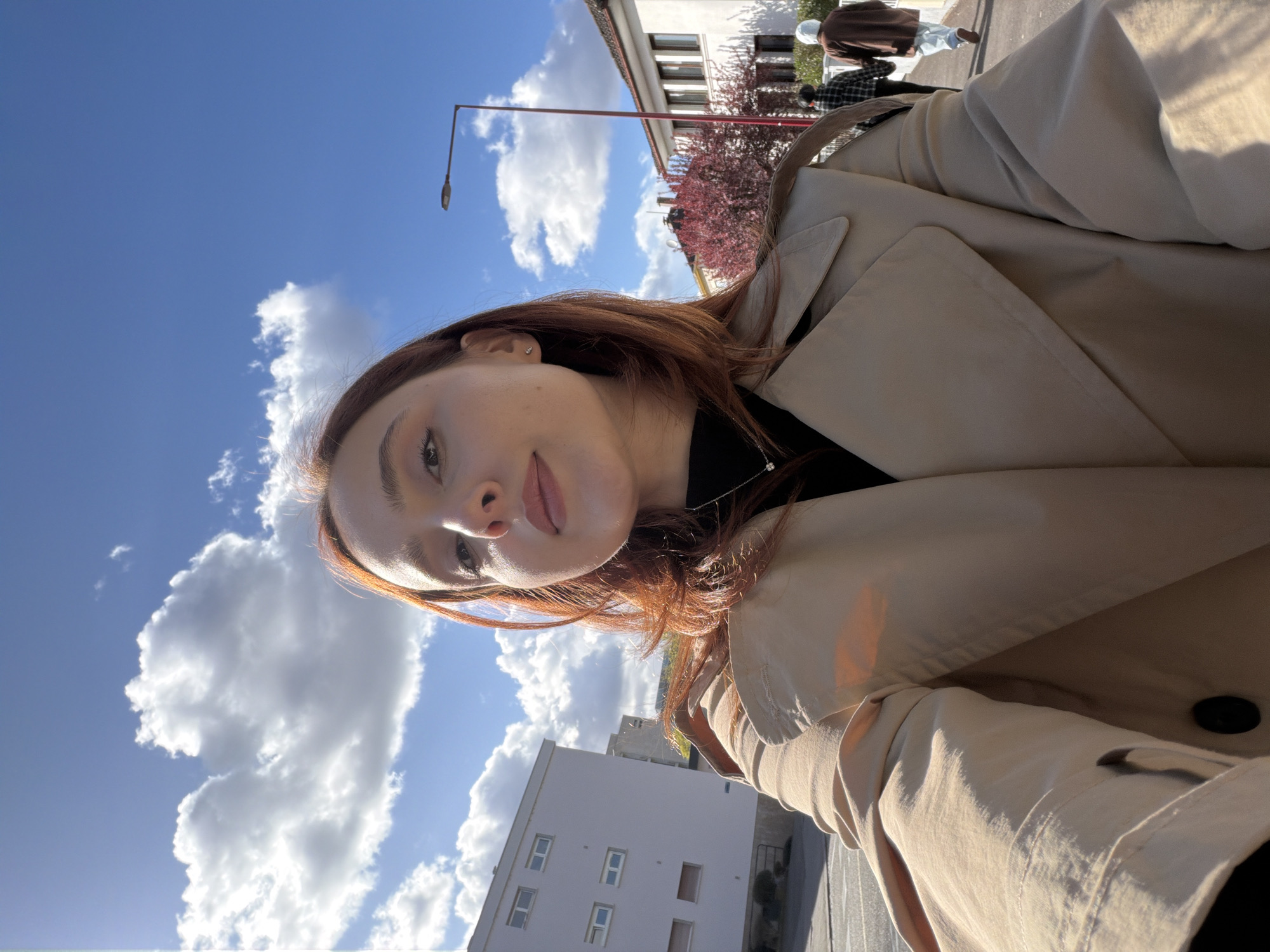 The image displays a selfie-style portrait of a young female outdoors in Strasbourg, France, under a clear blue sky with scattered white cumulus clouds. She has reddish-brown hair, light skin, and is wearing a black turtleneck shirt beneath a light beige trench coat. A silver necklace with a small pendant and a stud earring in her visible right ear are discernible. Her gaze is directed slightly upward toward the camera. Strong sunlight illuminates the scene, casting highlights on her and the environment. In the background, to the left, a modern, multi-story, light-colored residential building with rectangular windows is visible, overlooking a paved area. To the right, a sidewalk recedes into the distance where two individuals are walking away from the camera: one wears a dark checkered top and dark cap, the other a light brown long coat and a white head covering. A streetlamp with a red pole and a tree featuring vibrant red foliage are positioned along the path, in front of a white residential building with multiple windows.