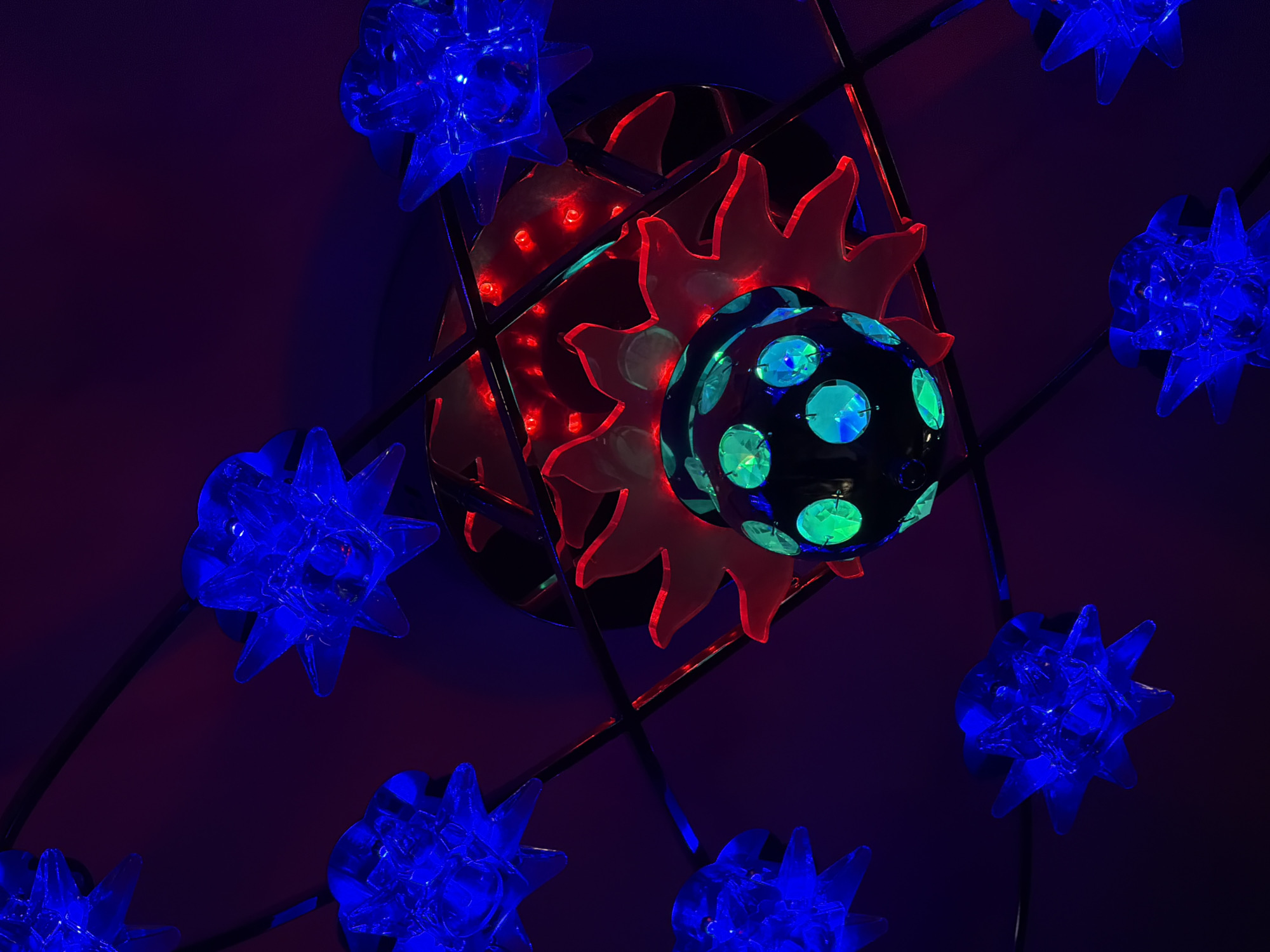 The image depicts a decorative light fixture, likely indoors due to the controlled lighting conditions. The fixture consists of multiple elements: several blue star-shaped lights are arranged around a central structure. At the center, a red sun-like shape appears, encircling a black sphere with green and blue reflective spots. Black lines form a grid connecting the elements. The background is a gradient of dark purple, further suggesting an interior space. The location is unknown.