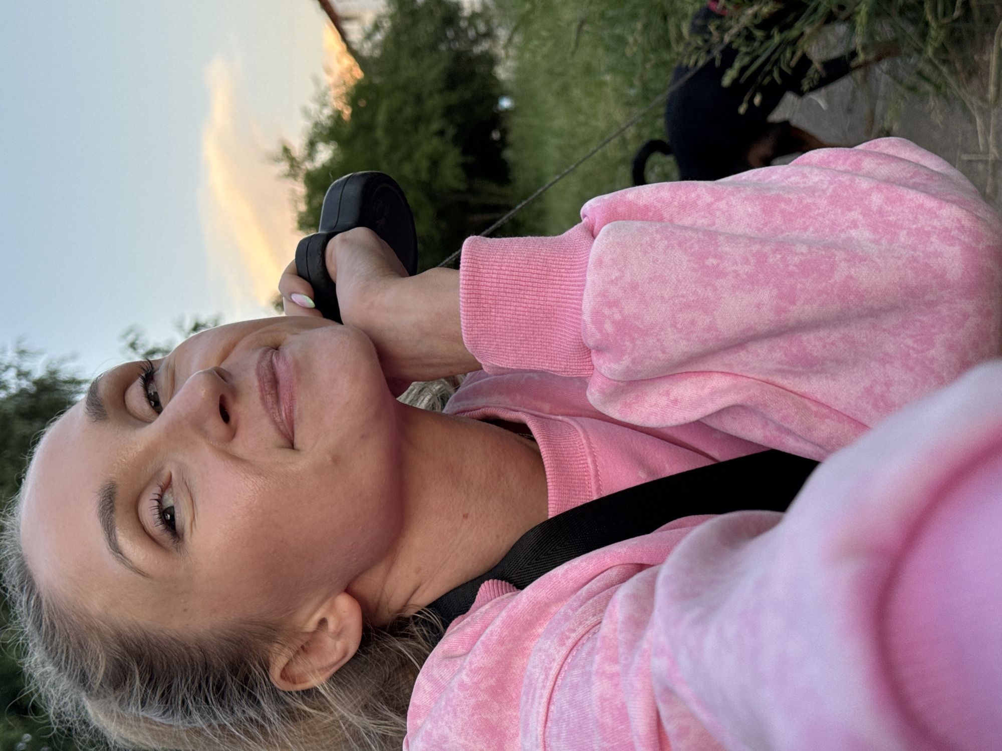 A close-up selfie captures a woman in a pink tie-dye sweatshirt holding a black object to her ear. Her left hand rests on her chin, and she is looking upwards with a gentle smile. Her blonde hair is pulled back into a ponytail. In the background, a dog on a leash is visible walking on a paved path through a grassy area with trees and a building. The sky is a soft blue with hints of orange clouds, suggesting it is sunset. The location is identified as Chaikovsky, Russia.
