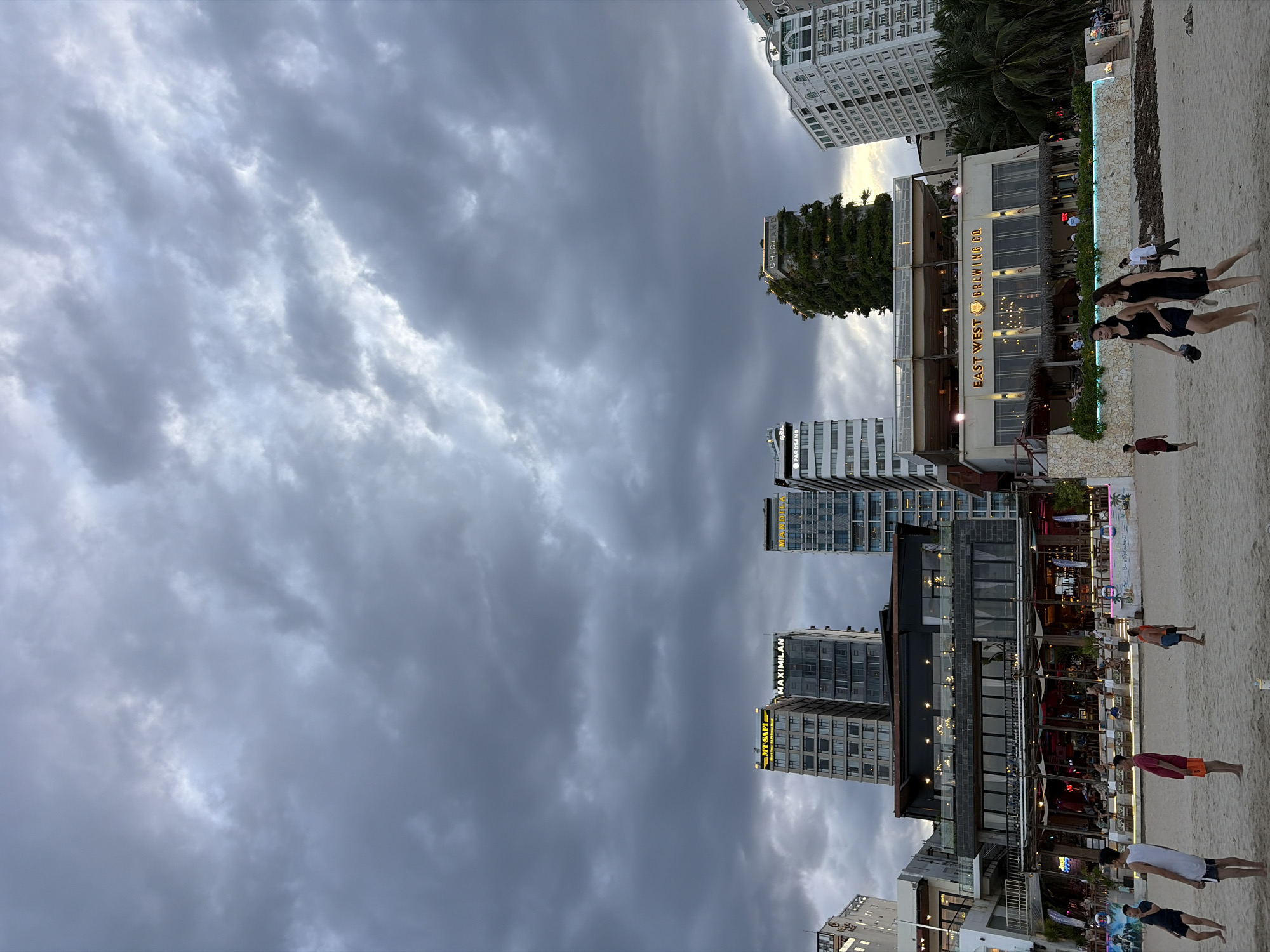 The image shows a cloudy sky over a beach scene in Đà Nẵng, Vietnam. In the foreground, several people are walking or standing on the sand. In the background, there is a row of modern buildings, including high-rise hotels and restaurants. One building prominently displays the text "EAST WEST BREWING CO." A lush green, tree-covered building stands out among the other structures. The beach appears to be a public space with some areas set up for dining or entertainment. The overall atmosphere is overcast but active.