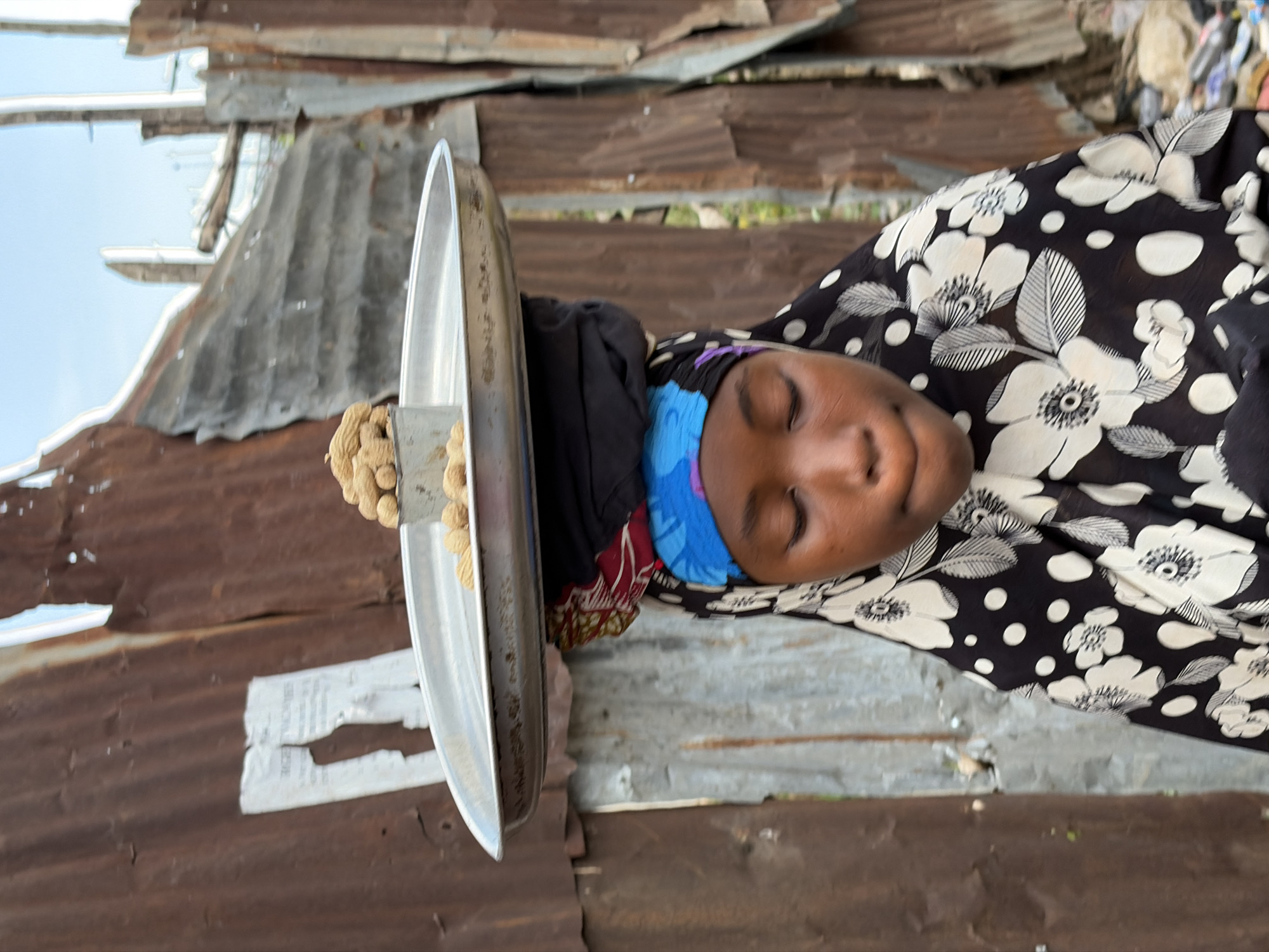 A woman wearing a headscarf and a black floral print dress is holding a silver tray with what appears to be groundnuts or peanuts on a small platform attached to the tray. She is in what seems to be a makeshift structure with walls and roofs made of corrugated metal sheets. The location is indicated to be Jos, Nigeria. The woman has her eyes closed, possibly resting or napping while waiting for customers.