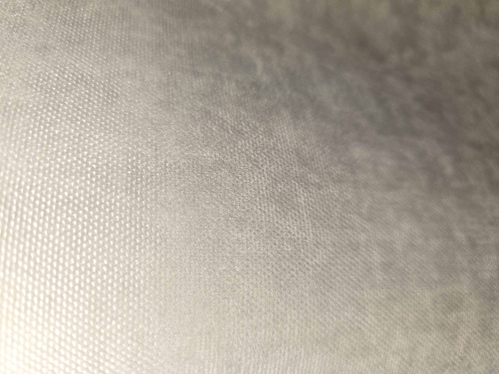 The image is a close-up, abstract shot of a textured surface. The texture appears to be a fine, raised pattern, possibly a fabric or a textured plastic, with small, repeating raised dots or bumps. The color is a light, neutral tone, likely a pale gray or off-white, with subtle variations in shading due to the lighting and the texture itself. The top left of the image is brighter, suggesting a light source from that direction, creating highlights and shadows that emphasize the texture. The focus is shallow, with the foreground texture being more distinct than the background, which becomes progressively blurred and darker towards the bottom and right of the frame. There are no discernible objects, people, or environmental context to determine a specific location. This image does not provide any information that can be used to determine its location in Ulan-Ude, Russia.