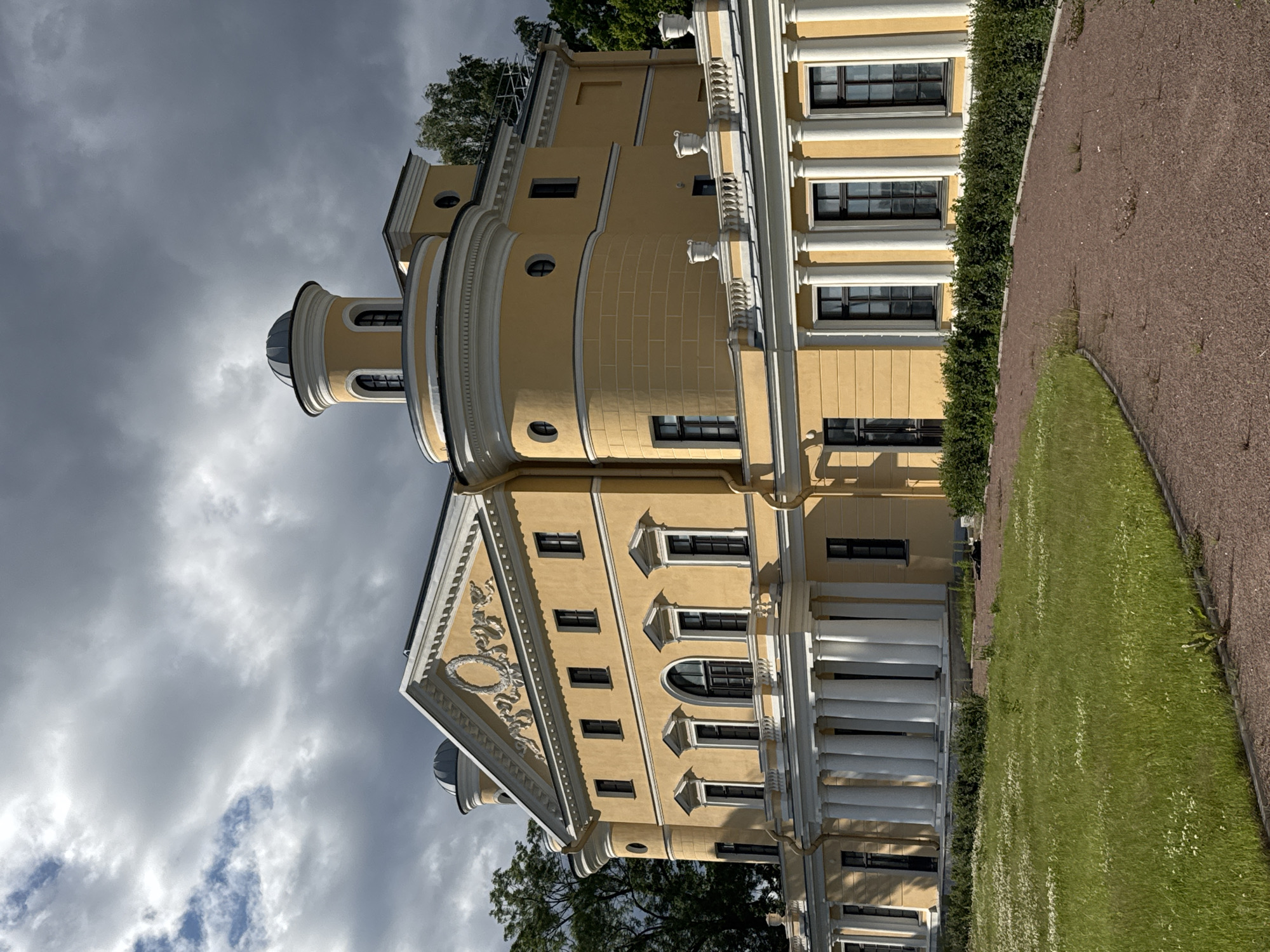 This is a medium-angle, full shot of a yellow neoclassical building, viewed from its side and below, with the camera rotated 90 degrees counterclockwise. The building has a domed turret and a classical facade with columns and decorative elements. A manicured lawn and a gravel path are visible in the foreground, bordered by a dense hedge. Trees are visible behind the building. The sky is overcast with dark clouds, suggesting a cool or inclement day, possibly late afternoon. The image is likely taken in Saint Petersburg, Russia, given the architectural style and mention of its location. No people or text are visible in the image.