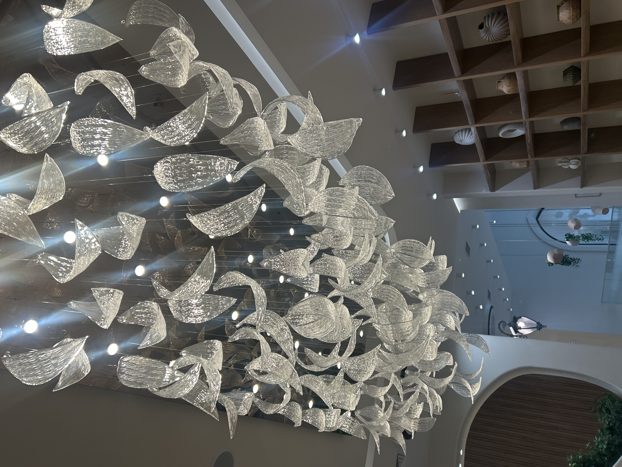 The image depicts a modern interior space, likely a commercial establishment or public area, featuring a large, elaborate chandelier made of clear, textured glass elements suspended from the ceiling. The chandelier's design is organic, with many individual pieces resembling flowing petals or leaves. Below the chandelier, to the right, is a tall, wooden shelving unit filled with various decorative ceramic vases and objects. On the left side, a curved architectural element with a wood-paneled surface is visible. Further in the background, a vintage-style lantern is suspended, and there are hints of other decorative elements and lighting fixtures. The overall impression is one of contemporary design with artistic touches. This scene is located in the city of Almaty, Kazakhstan.