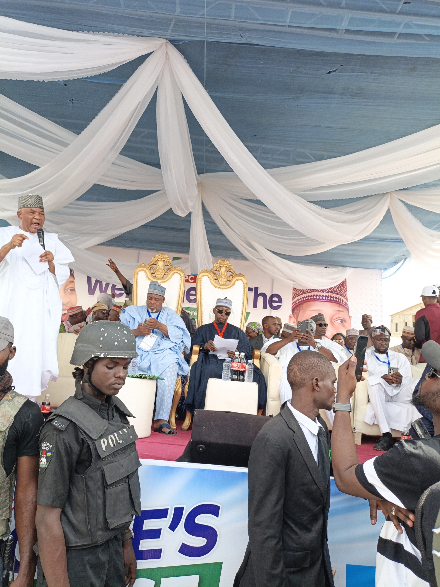 The image shows a political event taking place in what appears to be Giginyu, Nigeria. A stage is set up, draped with white fabric. On the stage, two men sit in gold-colored chairs, and a third man stands at a podium, speaking into a microphone and holding papers. The audience is visible in front of the stage, with many people holding up cellphones to take pictures or record videos. A uniformed police officer stands guard in the foreground. Bottles of Coca-Cola are visible on the stage beside the seated men.
